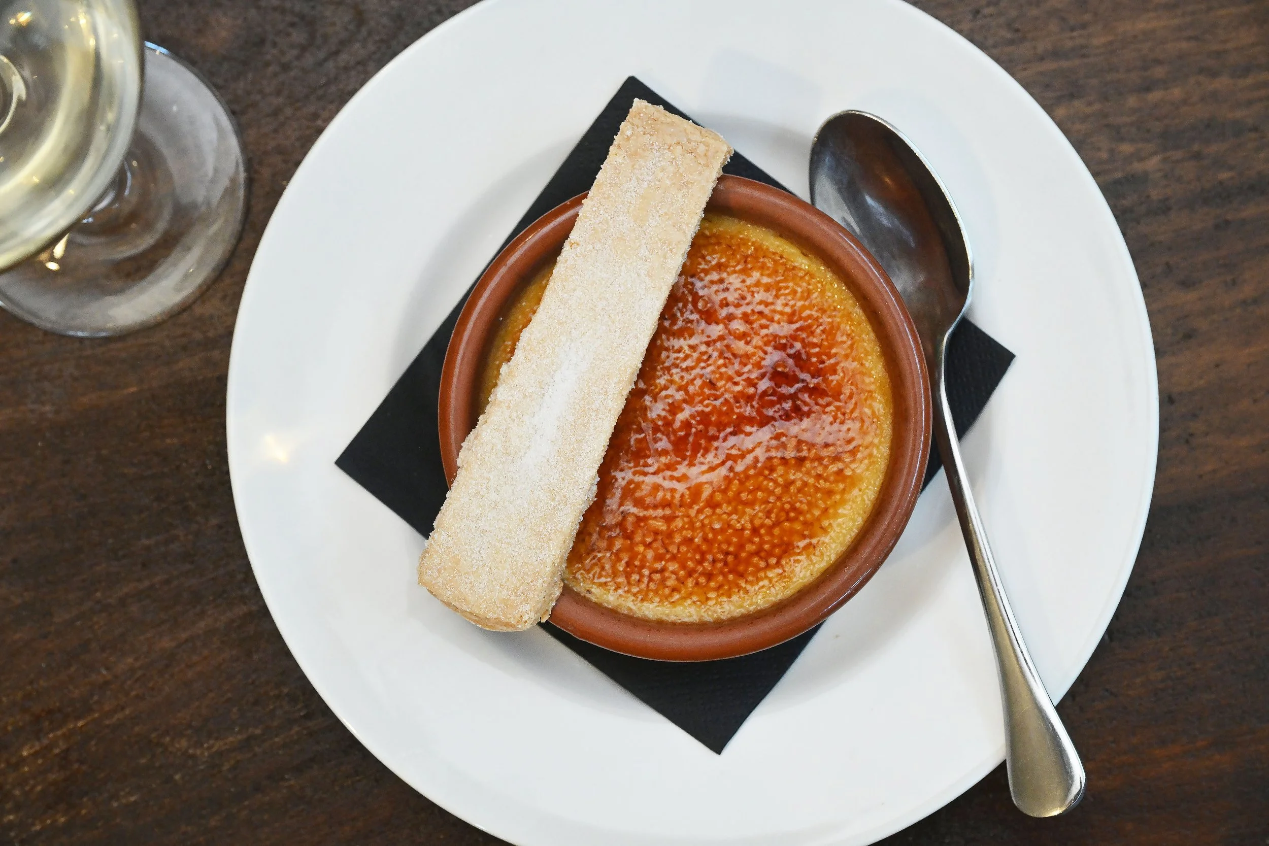 Crème brûlée in a small terracotta dish with a caramelized sugar top, garnished with a long, rectangular cookie on a white plate with a silver spoon, set on a dark wooden table, with a glass of white wine visible in the corner.