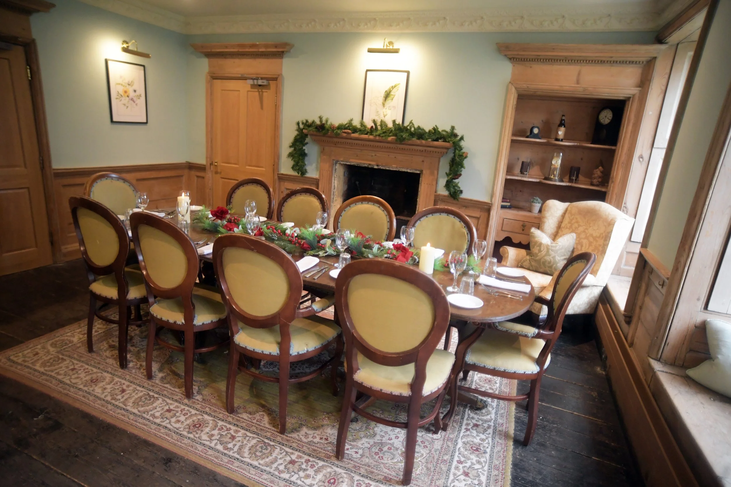 Decorated dining room with a long wooden table set for a festive meal, surrounded by upholstered chairs, a fireplace with a mantel decorated with greenery, and a window seat with cushions.