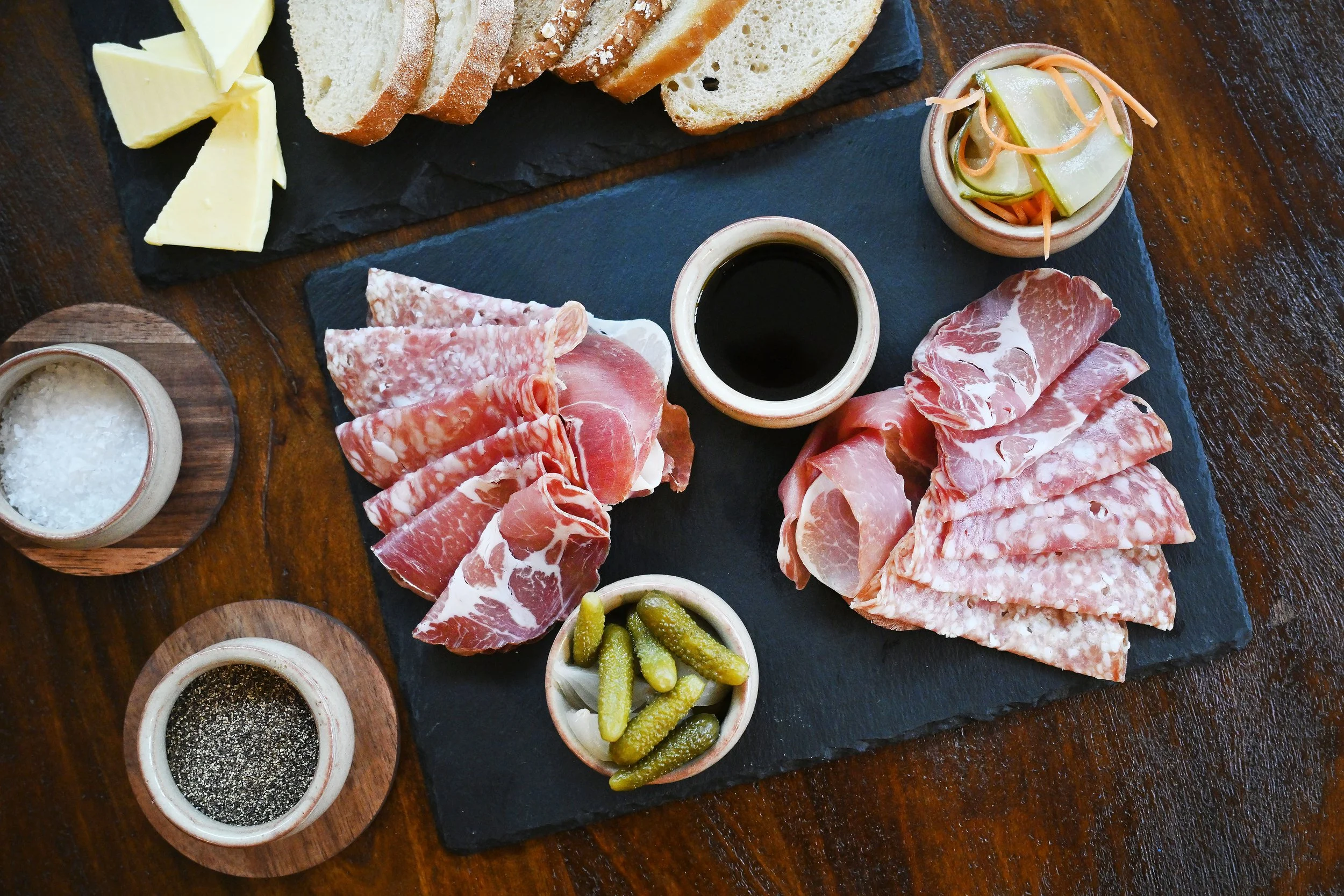 Assorted sliced cured meats, pickles, and sauces on a black slate serving board with bread and cheese in the background on a wooden table.