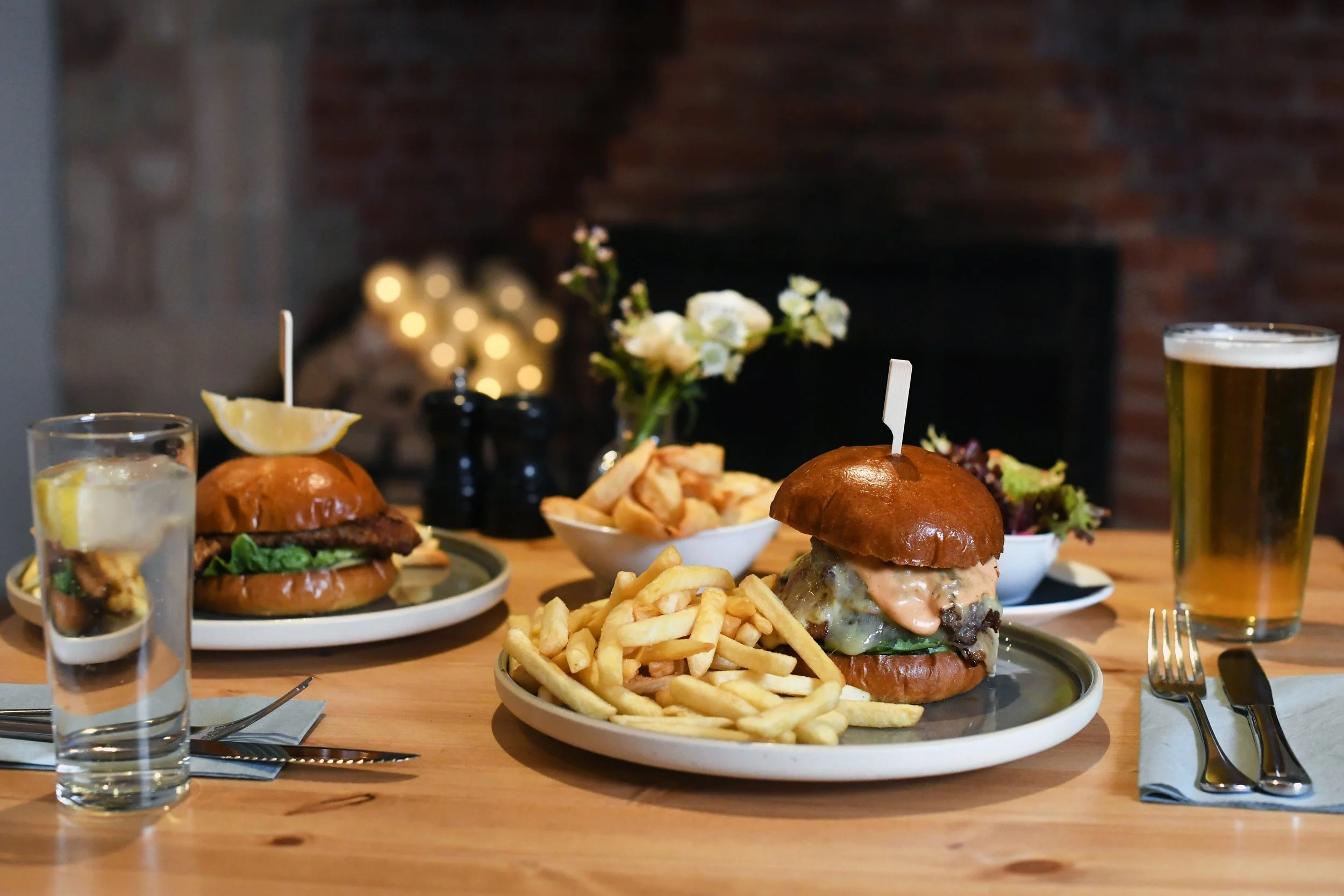 Two plates of one burger with lettuce, cheese, and sauce, French fries, a bowl of additional fries, a Salad, a glass of water with lemon, and a glass of beer on a wooden table, with a blurred background of a fireplace and decorative lights.