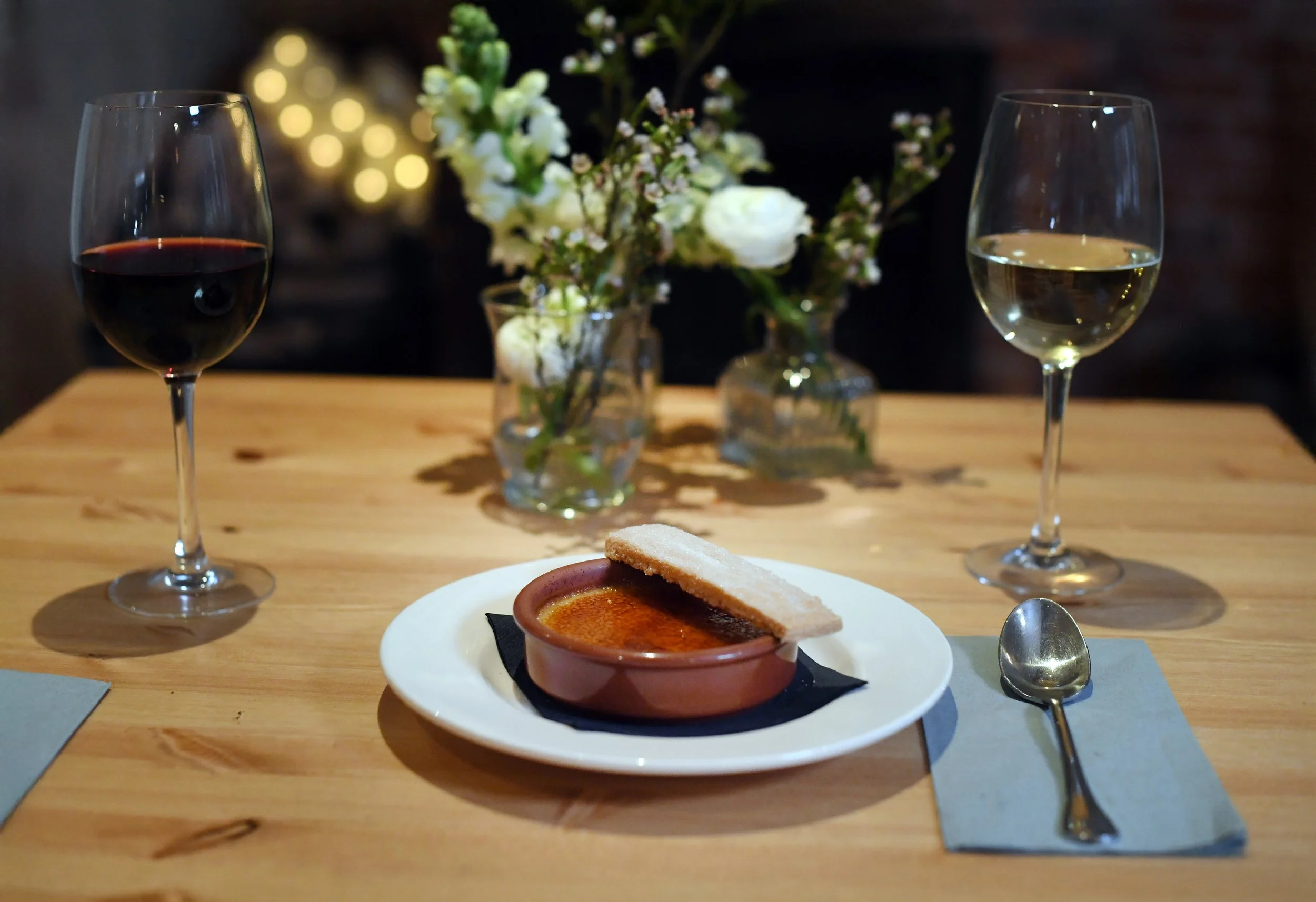 A dining table set with two glasses of red and white wine, a small plate with a crème brûlée topped with a cookie, a spoon on a napkin, and a floral centerpiece with white flowers in glass vases.