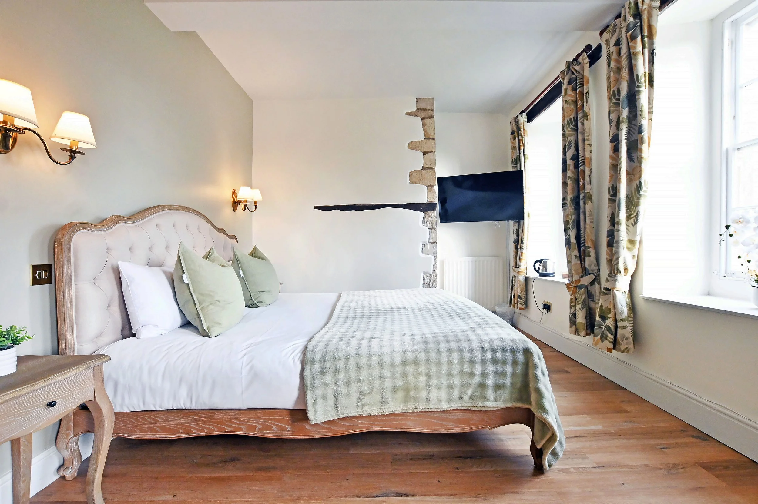 A bedroom with a large bed, light-colored tufted headboard, three green pillows, and a checkered blanket. There are wall-mounted lamps on a cream wall, a wooden side table with a potted plant, a flat-screen TV mounted near a window with floral curtai