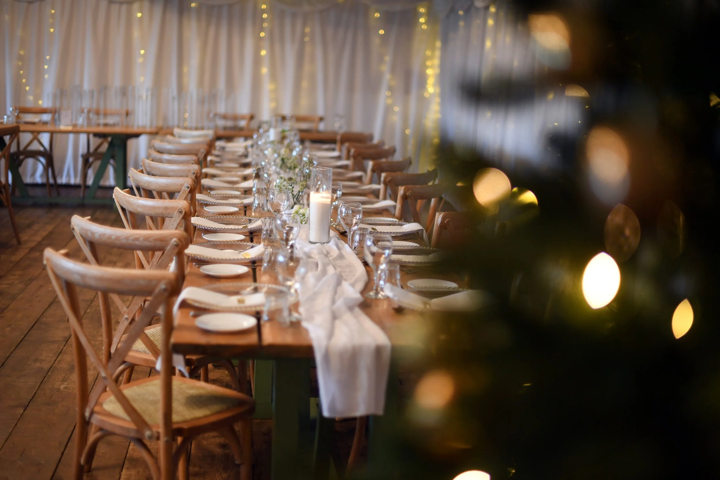 A decorated dining table set for a celebration with white plates, silverware, wine glasses, and a white table runner, illuminated by candles and fairy lights, with a white curtain backdrop.