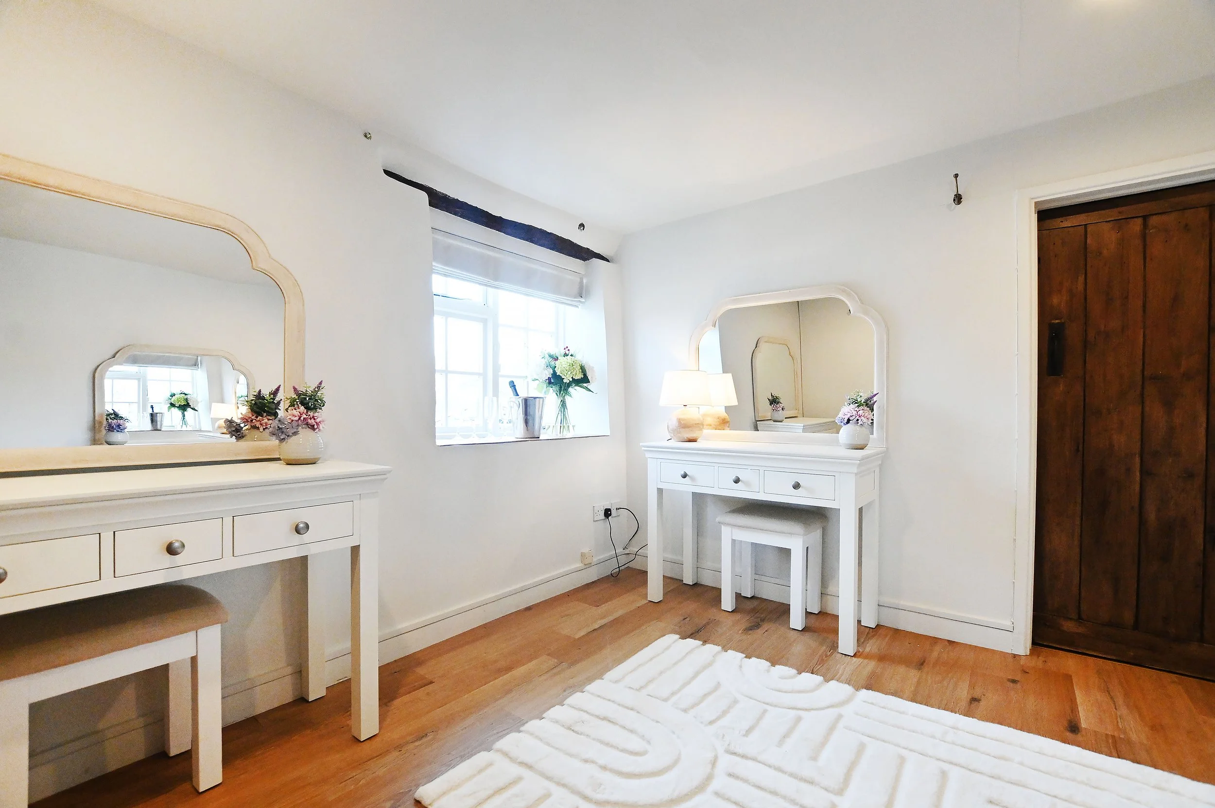 A bright bedroom with white furniture, including a dresser with a mirror, another smaller dresser with a mirror, a bench, and floral arrangements in vases. There is a window with a white blinds, a lamp, and a wooden door on the right. A white textured rug is on the wooden floor.