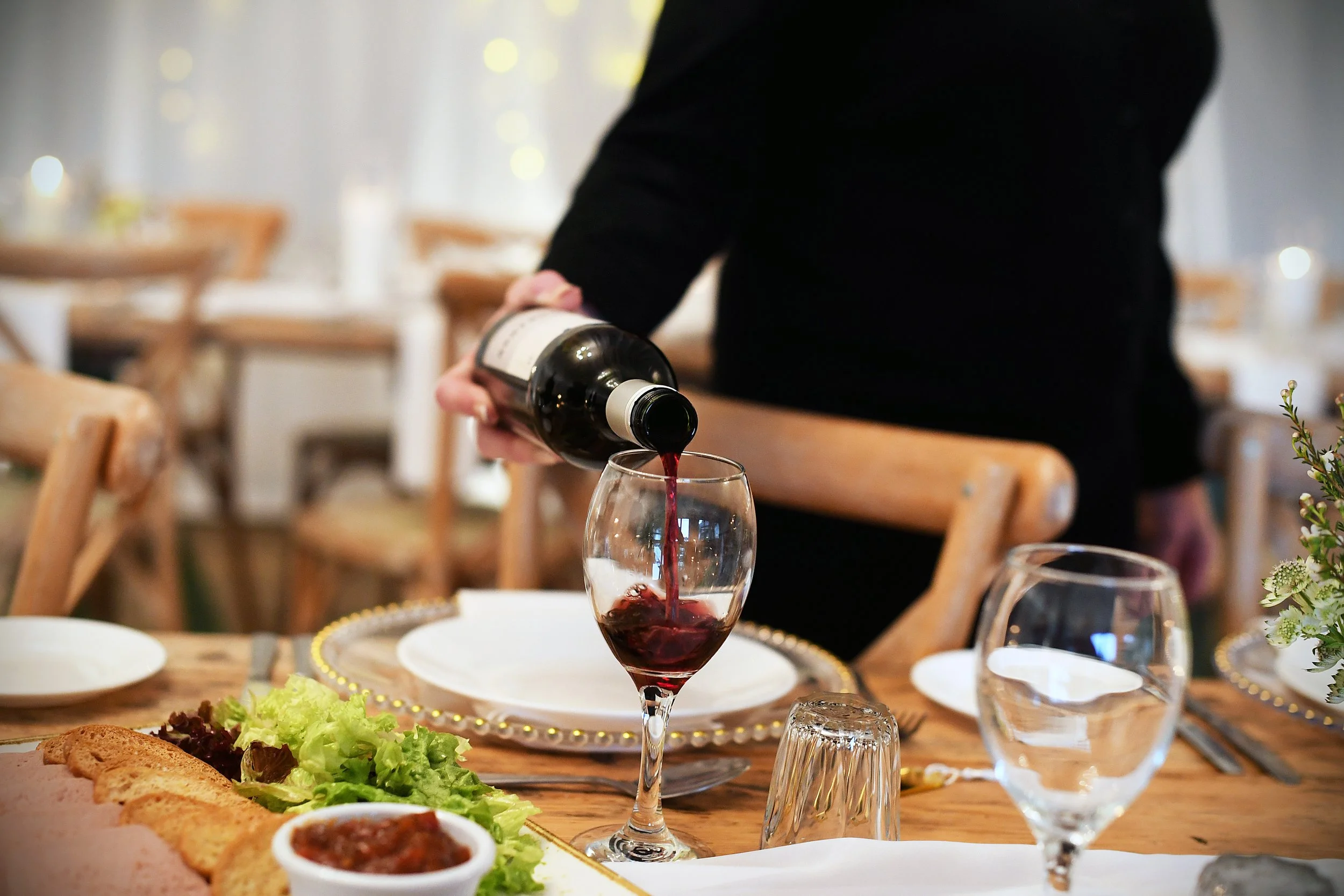 Person pouring red wine into a glass at a dining table set for a meal.
