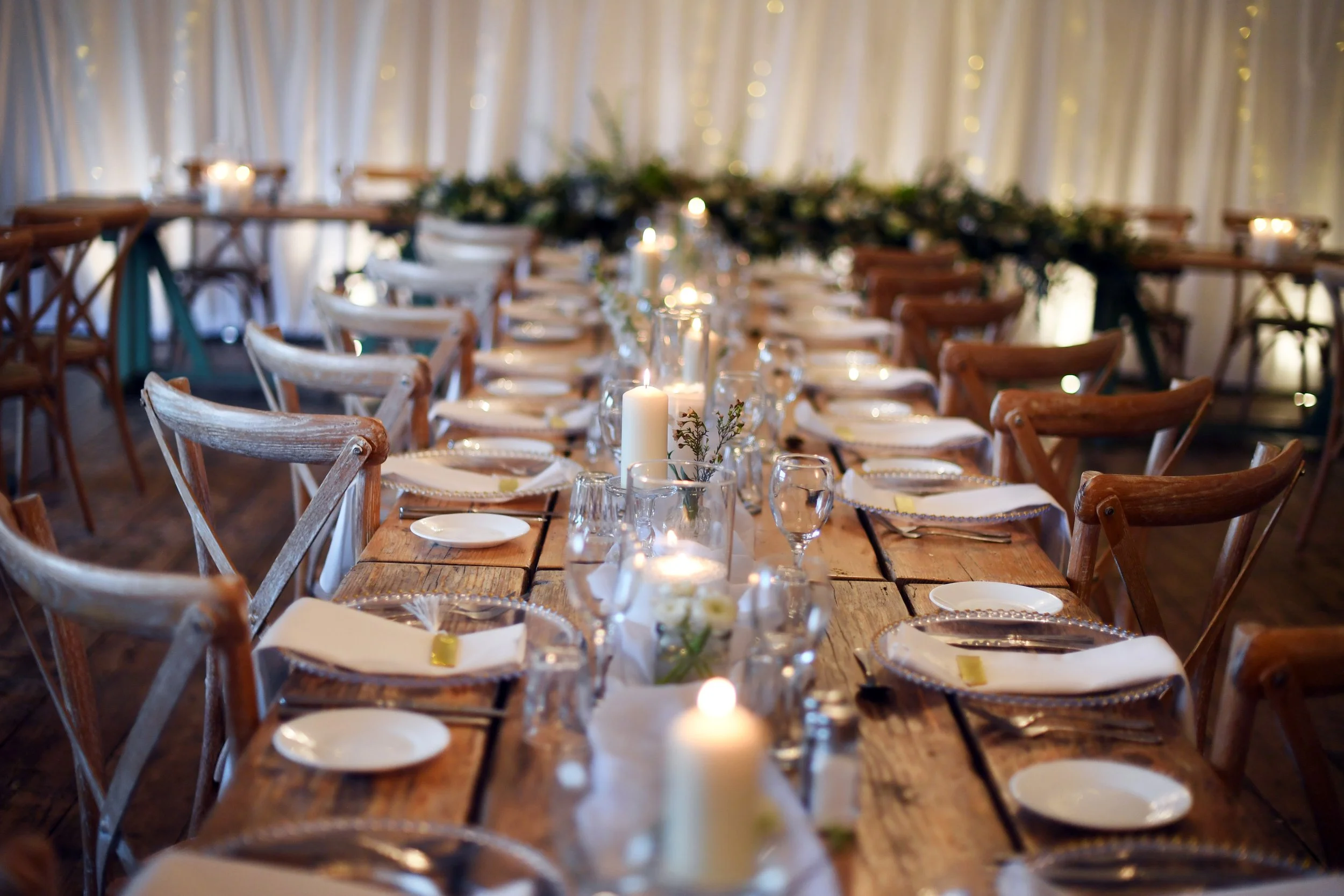 Long wooden dining table set with plates, glasses, napkins, and candles in a decorated venue with draped curtains and string lights.
