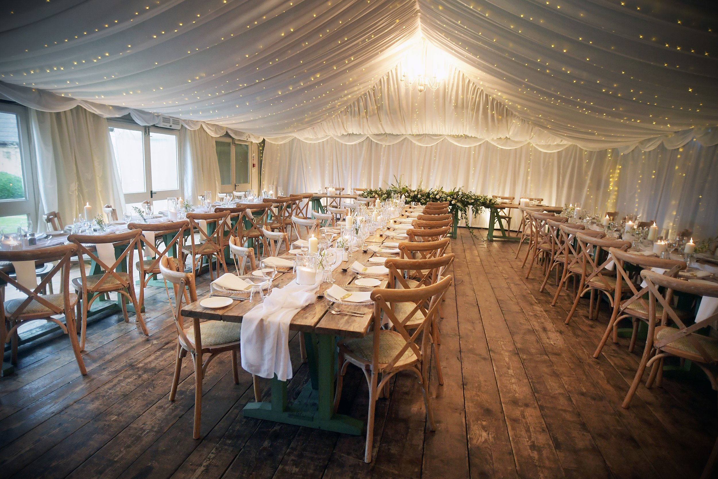 Elegant wedding reception tent with long wooden tables decorated with candles, white table settings, and greenery. Draped ceiling with fairy lights and a chandelier, rustic wooden floor, and windows on the side.