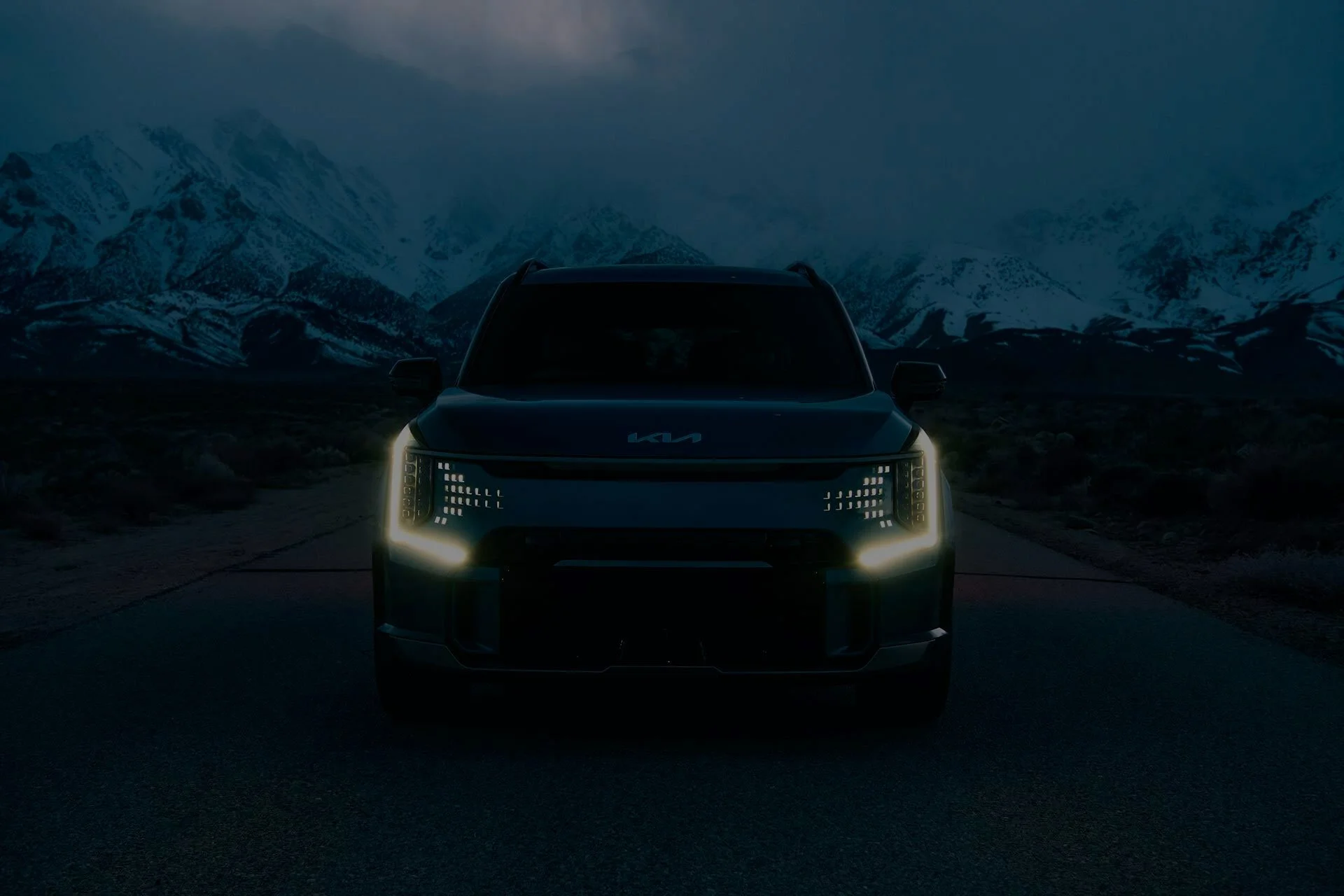 A dark Kia SUV with illuminated headlights parked on a road in a mountainous landscape at dusk or dawn.