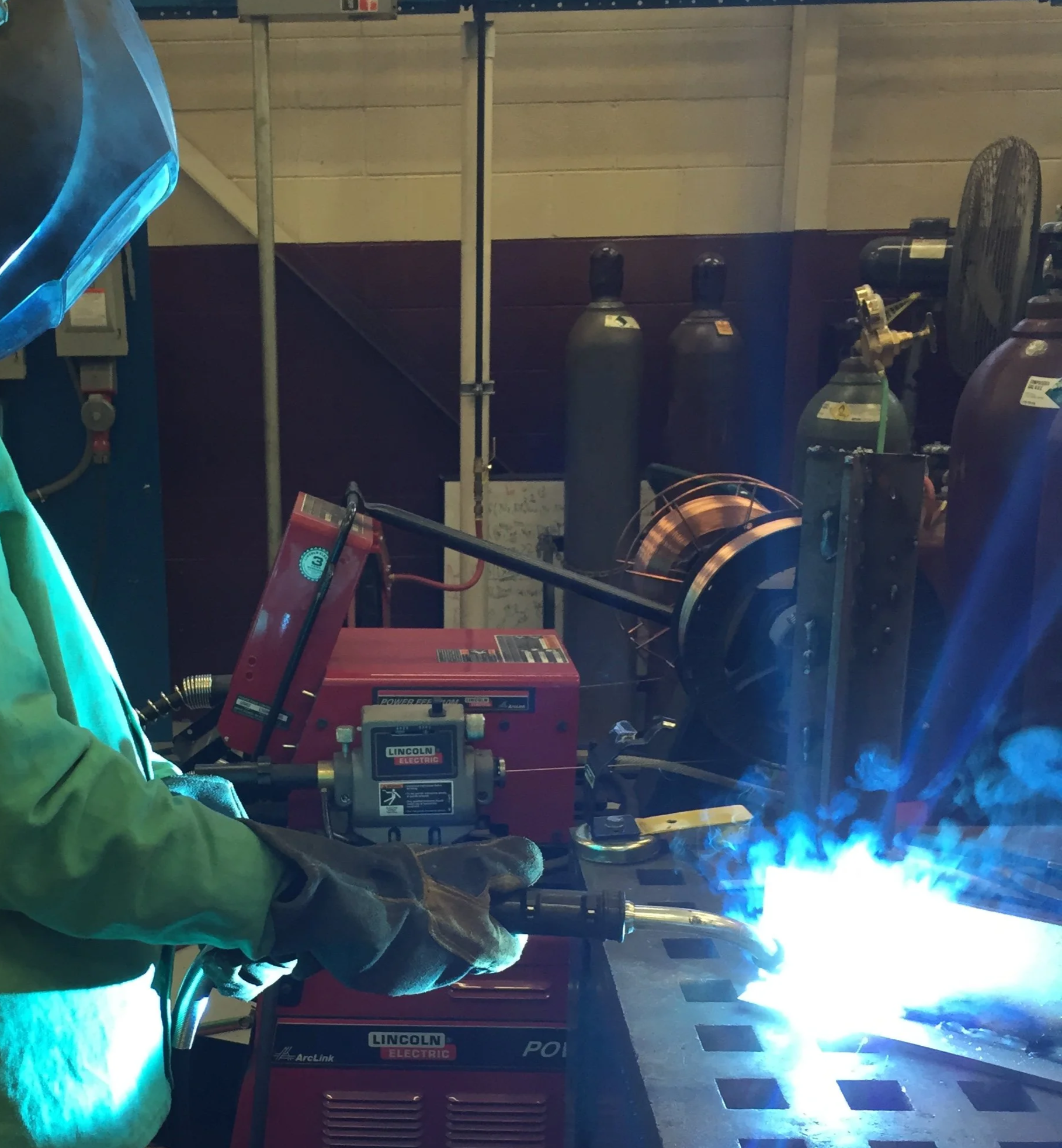 Worker welding metal on a workbench in a workshop, with welding equipment and gas cylinders in the background.