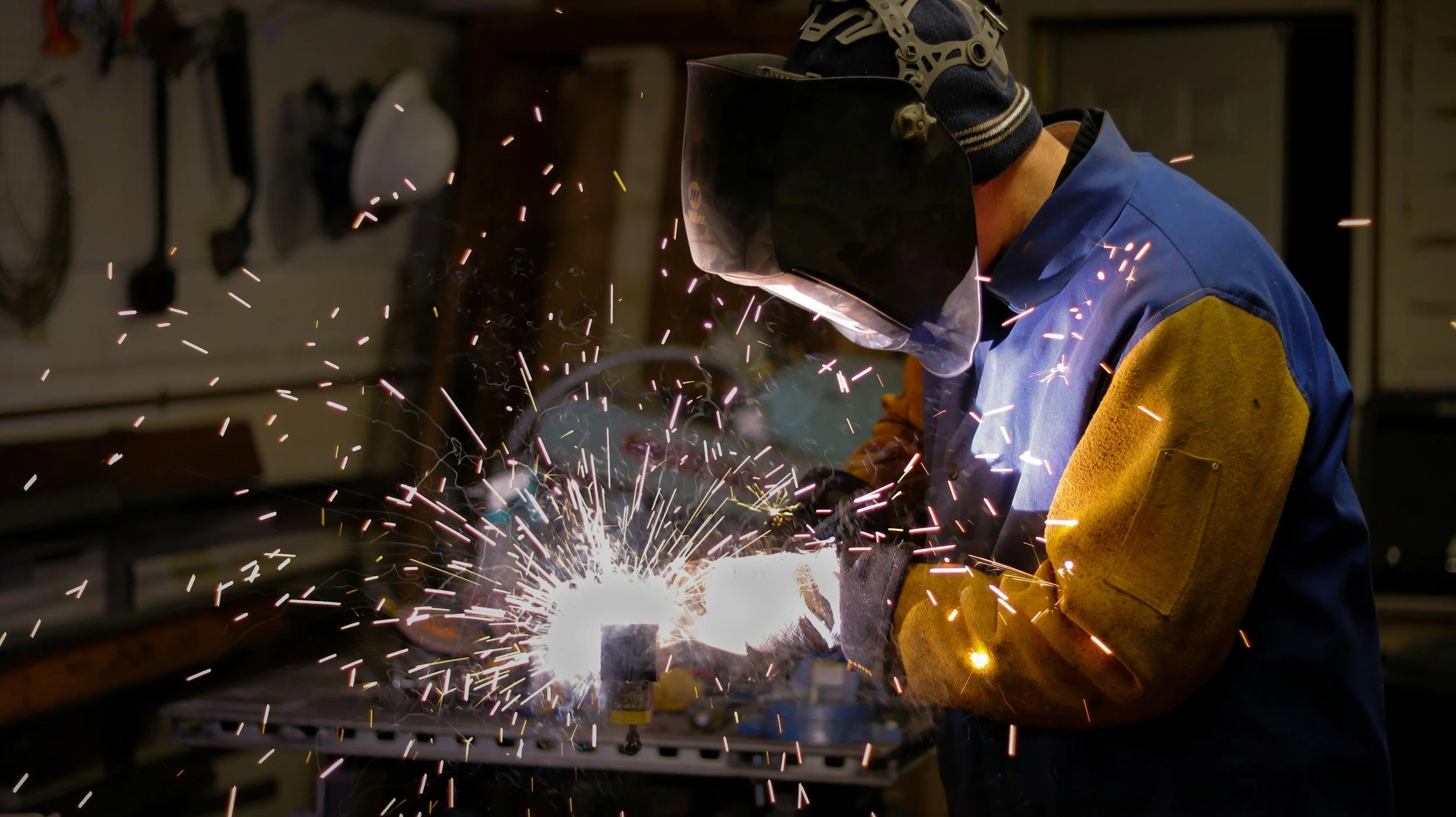 A welder wearing protective gear, including a helmet and gloves, welding metal producing sparks in a workshop.