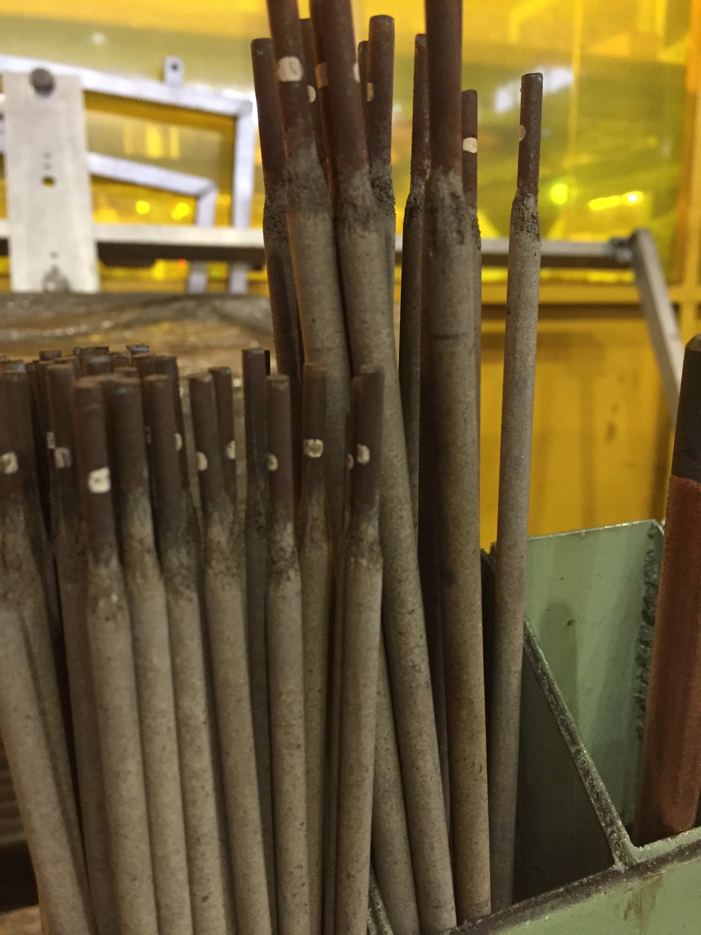 A pile of rusty metal rods standing vertically in a container, with some rods taller than others, against a yellow background.