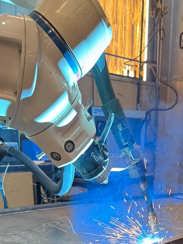 A robotic arm welding metal sheet with sparks and blue light in an industrial setting.