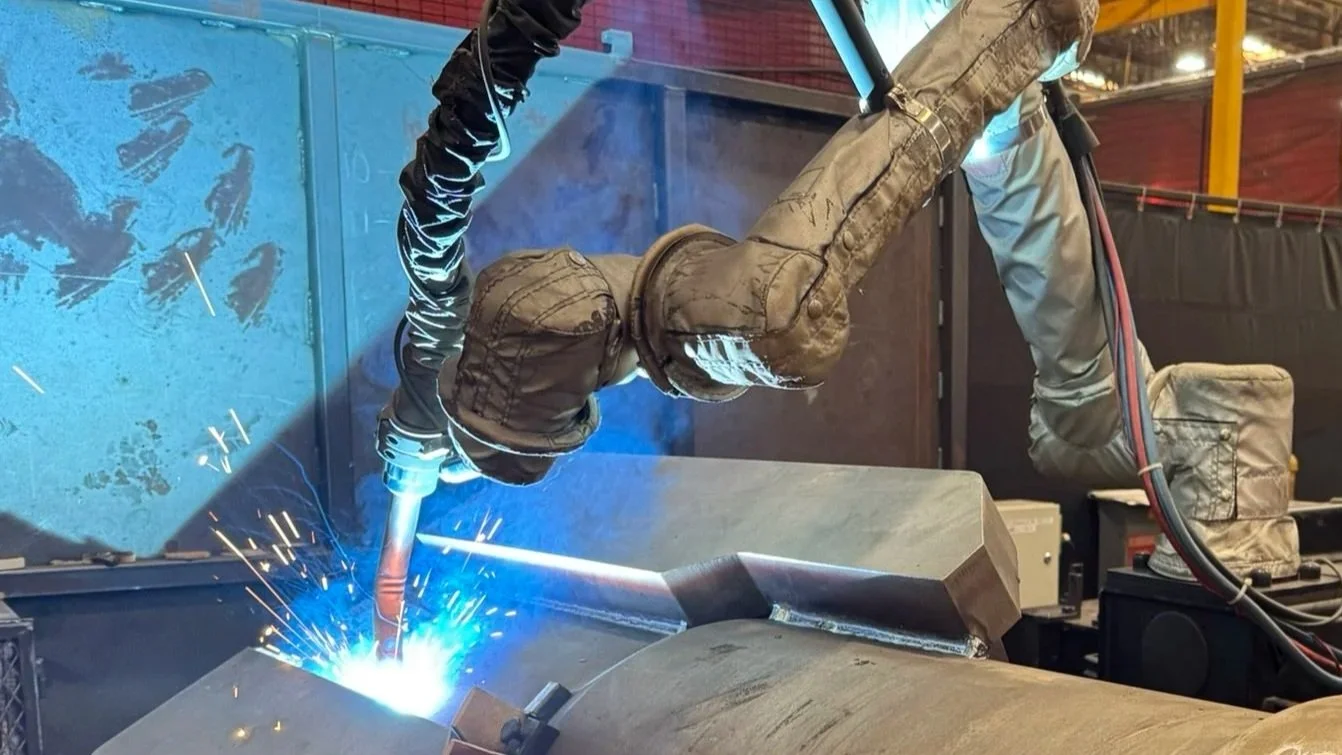 A person in protective gear is welding a metal piece on a large pipe, with sparks flying from the welding process.