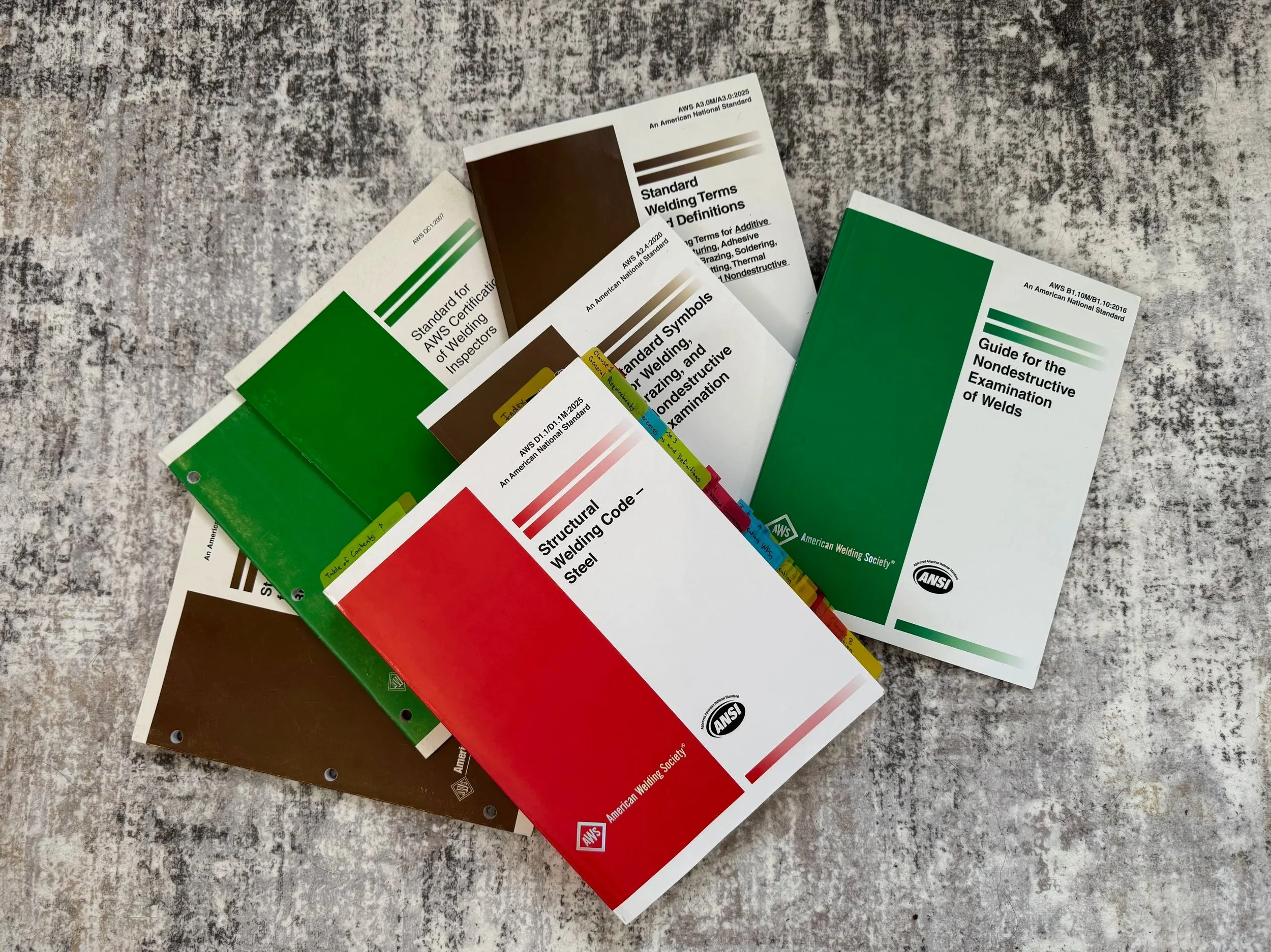 Several welding standard guides and codes lie on a textured gray surface. The guides feature various colors including white, green, brown, and red, each with titles related to welding terminology, inspection, and nondestructive examination.