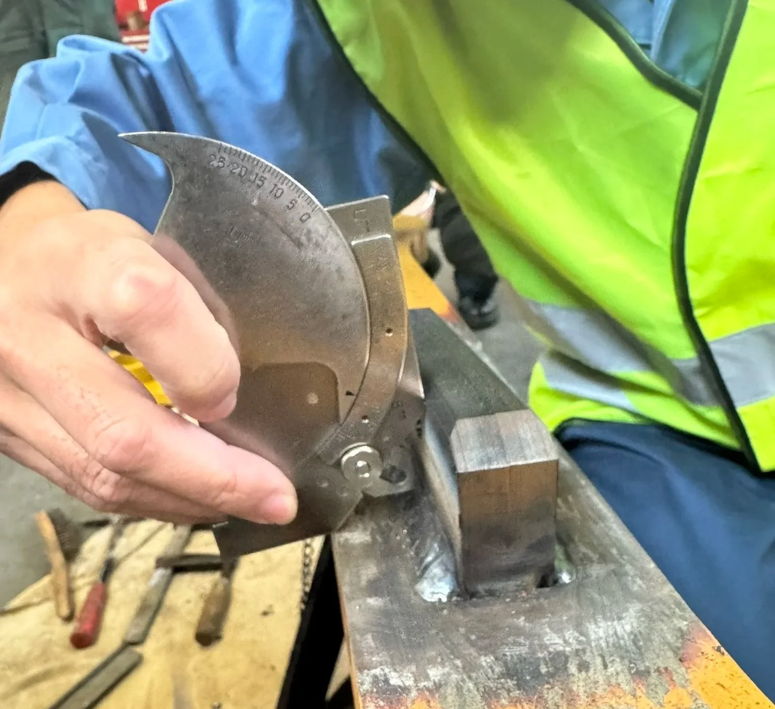 A person using a protractor to measure and mark metal on a workbench. The person is wearing a yellow reflective safety vest and a blue jacket.