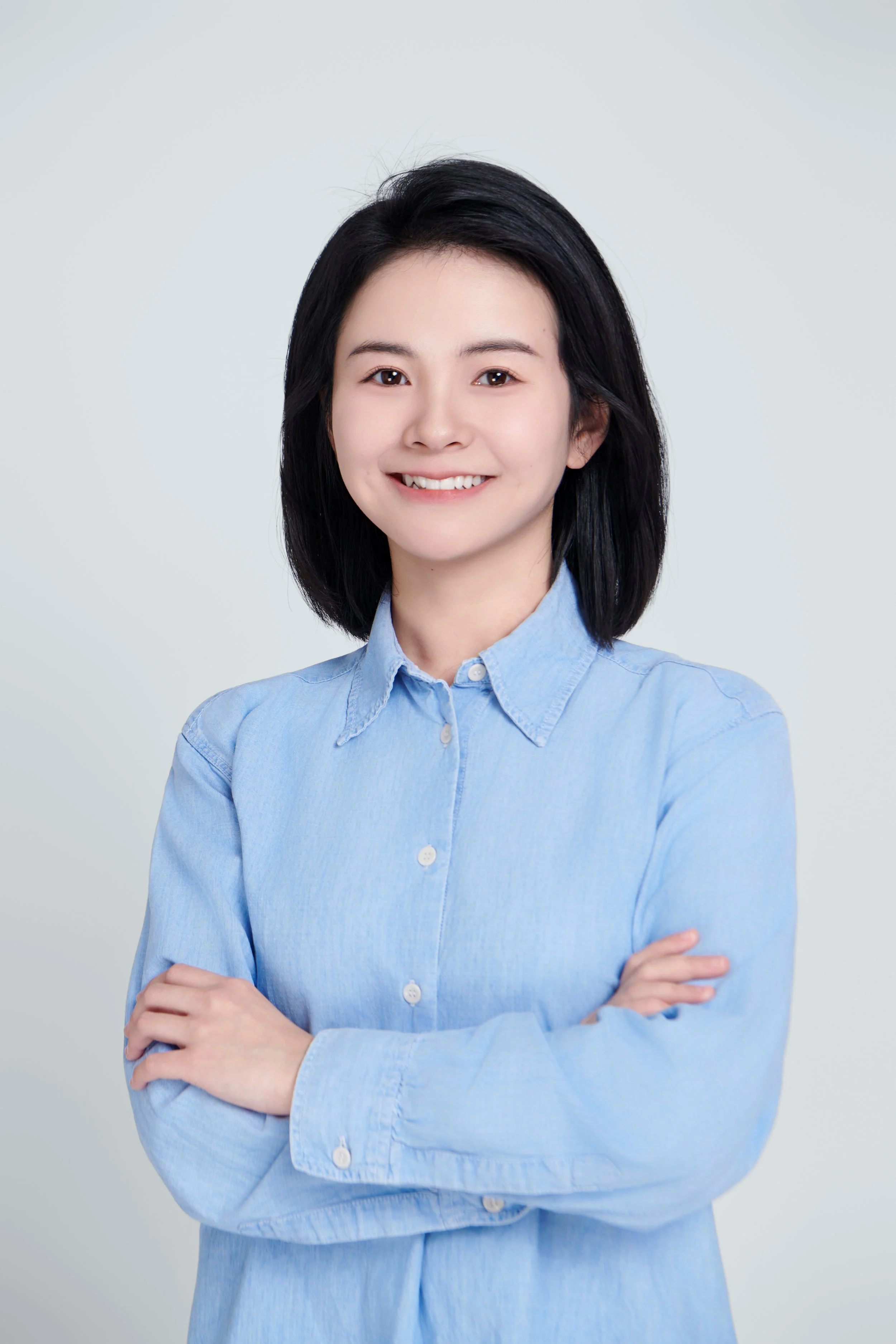A young woman with shoulder-length black hair, smiling, wearing a light blue button-up shirt, with arms crossed, standing against a plain light gray background.