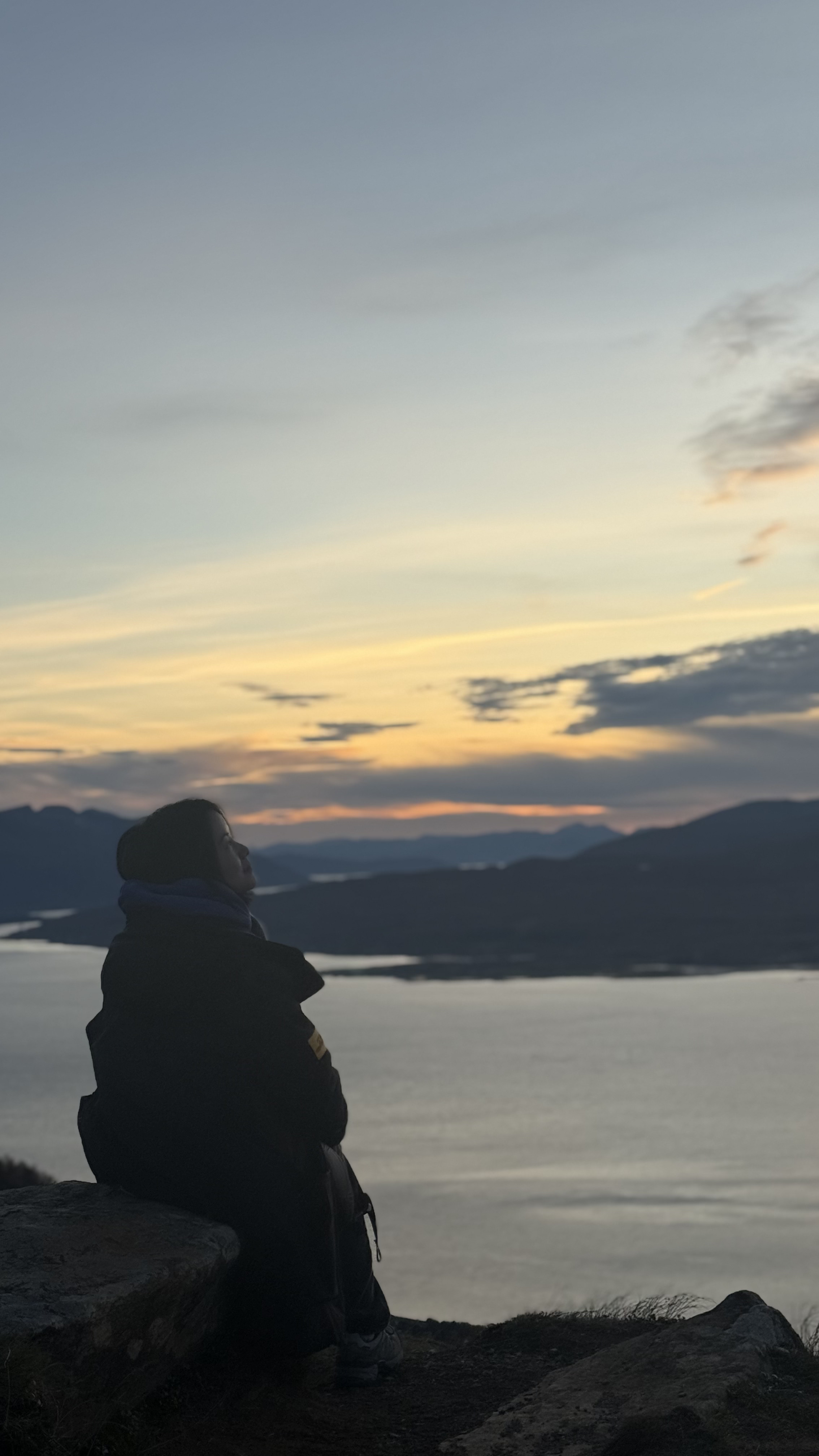 Person sitting on a rock overlooking a lake at sunset, with mountains in the background.