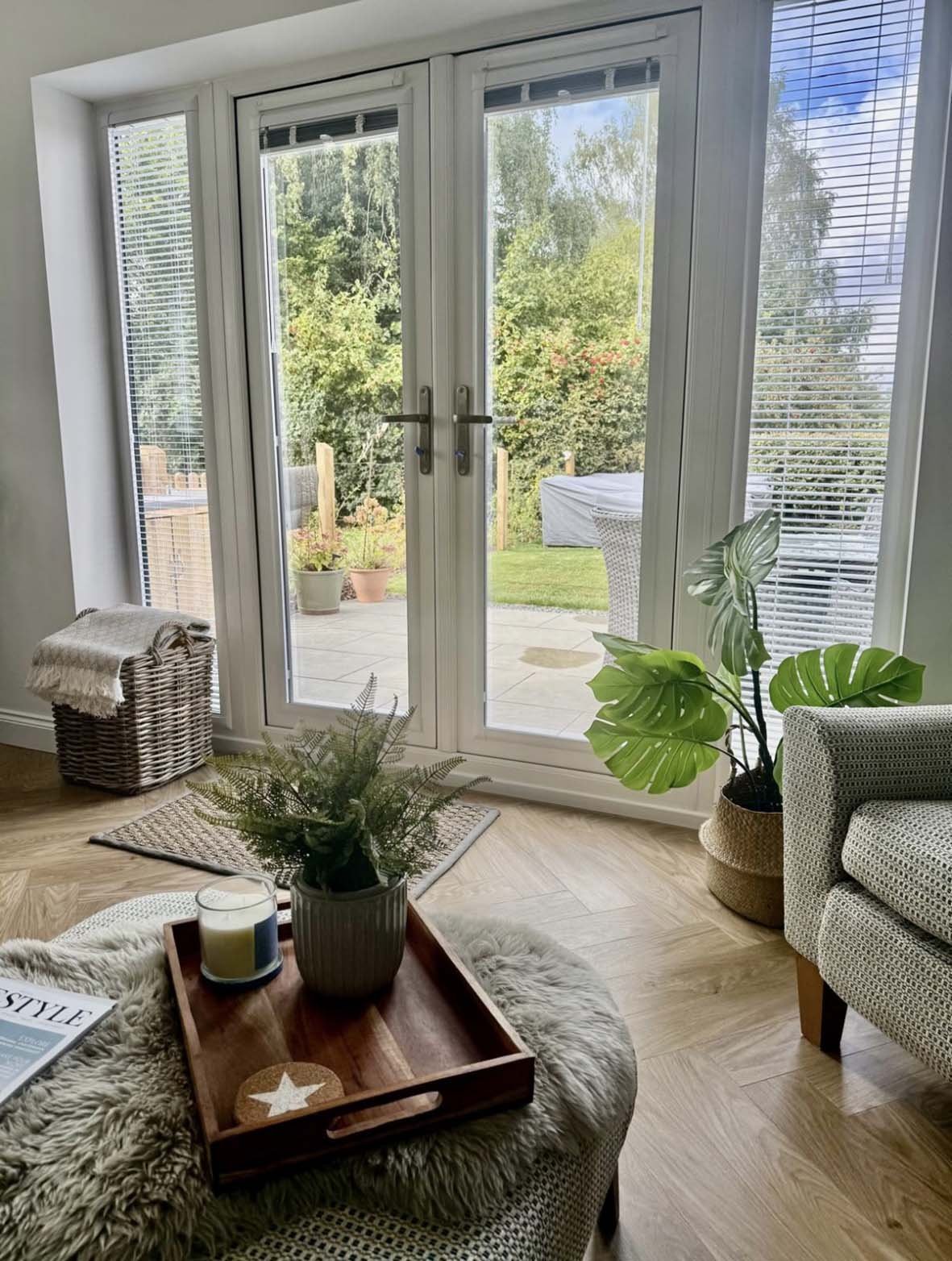 Living room with glass double doors leading outside, decorated with plants, a tray, candle, and magazine on a fluffy ottoman, and a patterned armchair nearby.