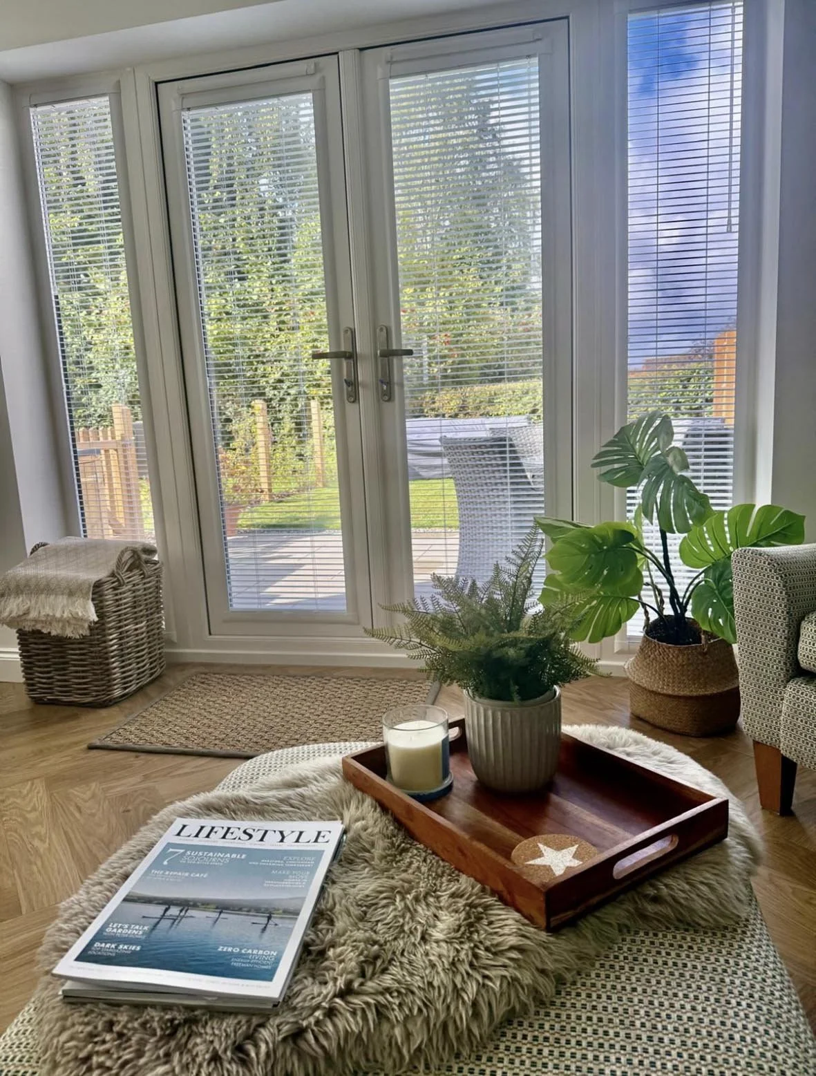 Living room with a large glass door leading to a garden, with white blinds. There is a wicker basket with a blanket, a small rug on wooden floor, a coffee table with a shiny fur blanket, a magazine, a tray with a potted plant and a candle, and a larg