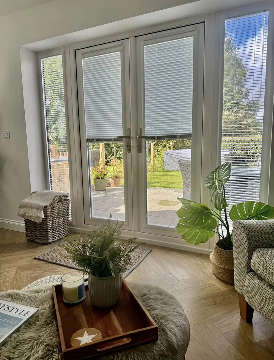 Interior view of a cozy living room with large glass doors leading outside, decorated with potted plants, a wicker basket with a blanket, and a small tray with candles on a plush rug.