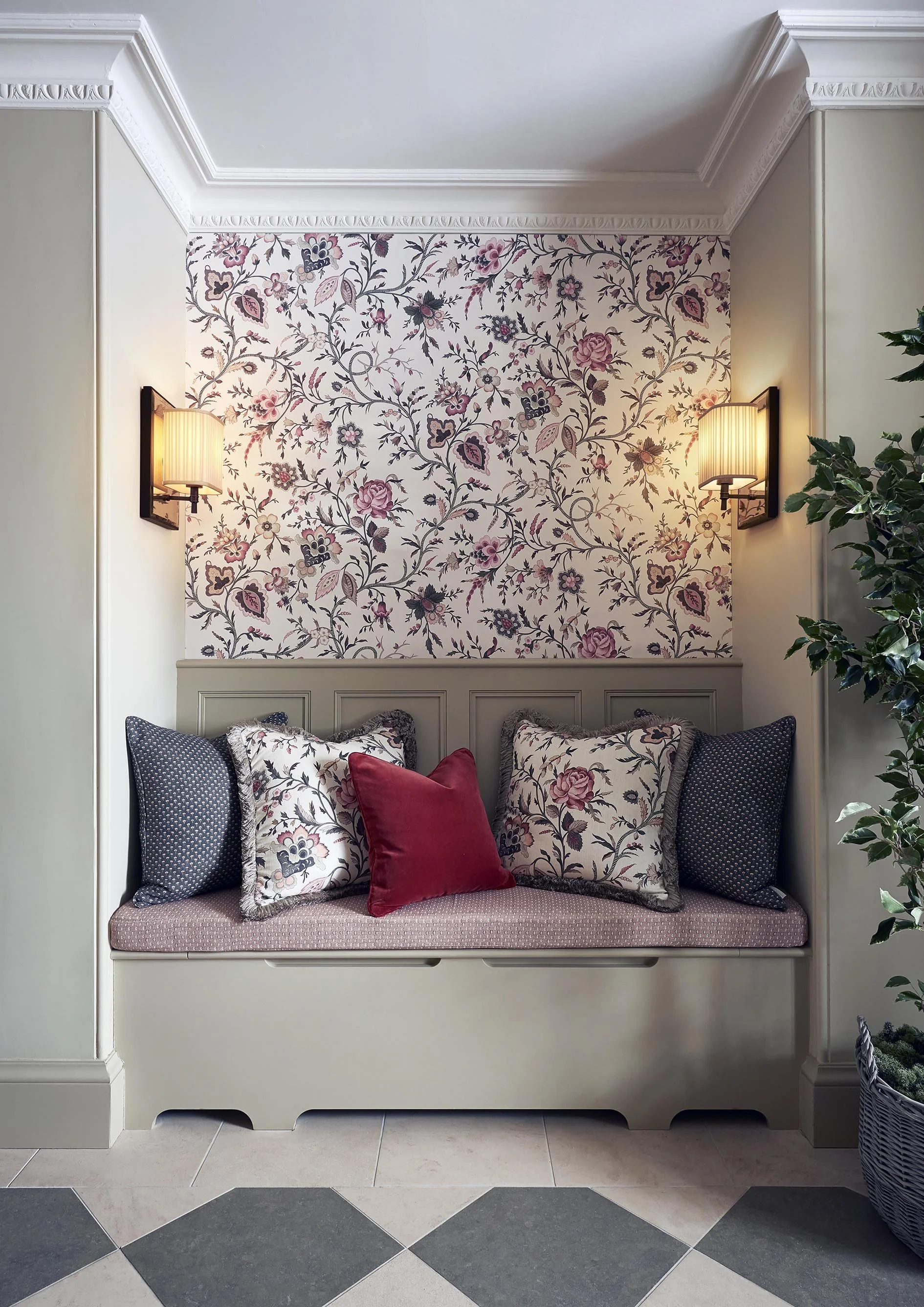 A cozy window seat with multiple decorative pillows, floral wallpaper on the wall behind, wall sconces on either side, and a potted plant on the right side.