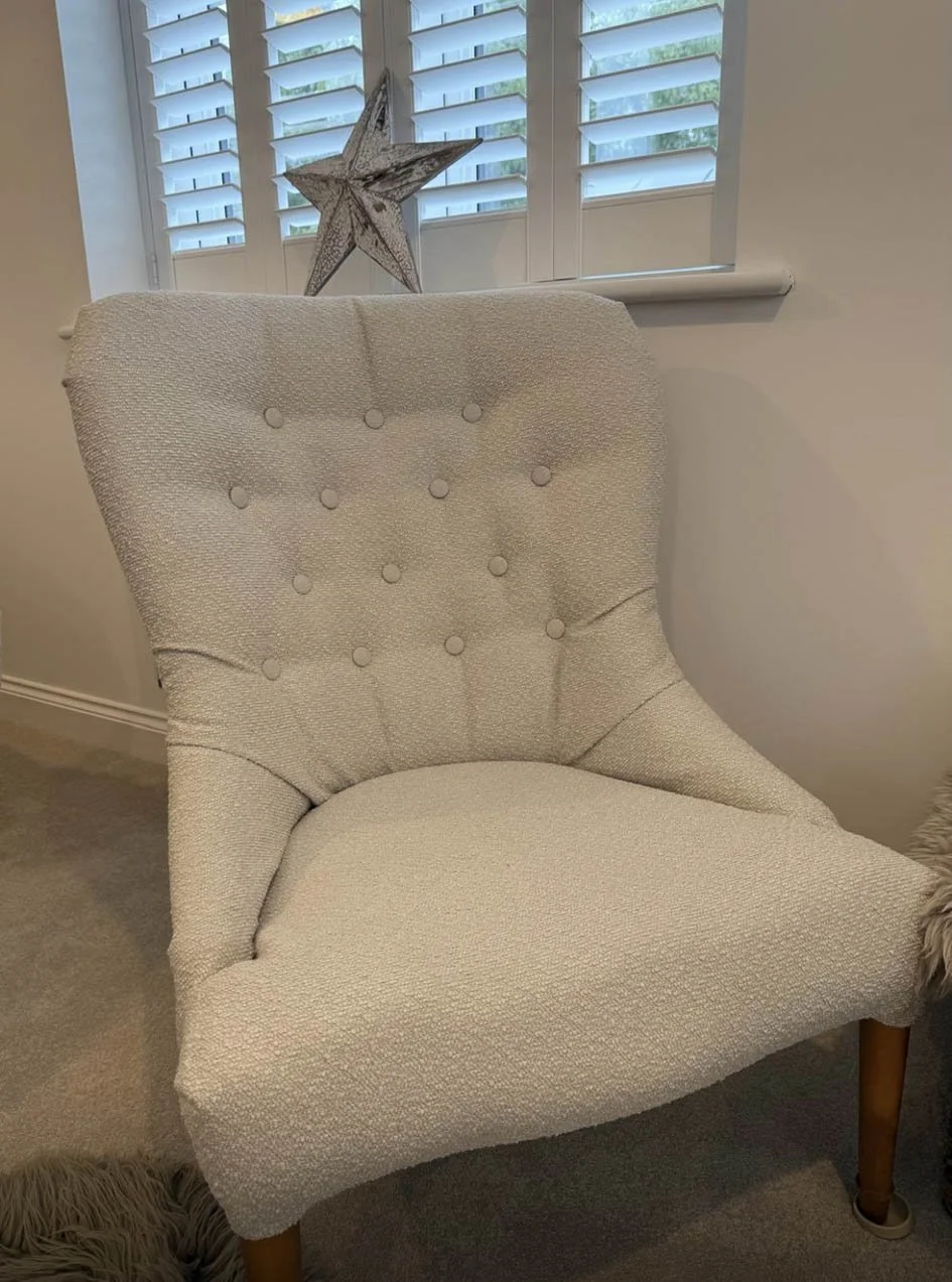 A cream-colored upholstered armchair with wooden legs, positioned in front of a window with white plantation shutters. A decorative star-shaped ornament is placed on the window sill behind the chair.
