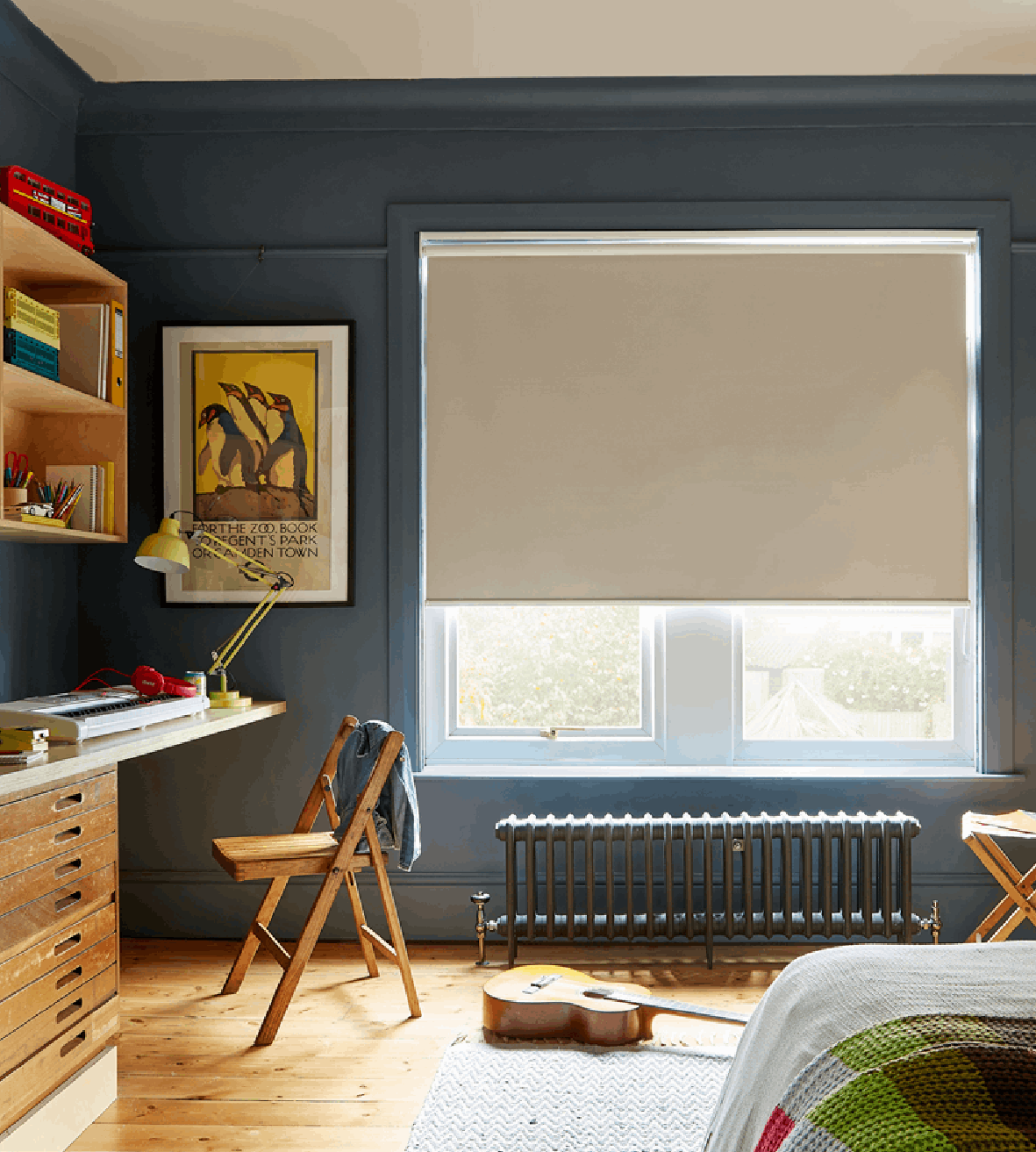 A cozy bedroom with a window covered by a beige roller blind, a wooden desk with a yellow task lamp, a wooden chair with a jacket draped over it, a framed art print of penguins, a guitar on the floor, and a bed with a colorful blanket.