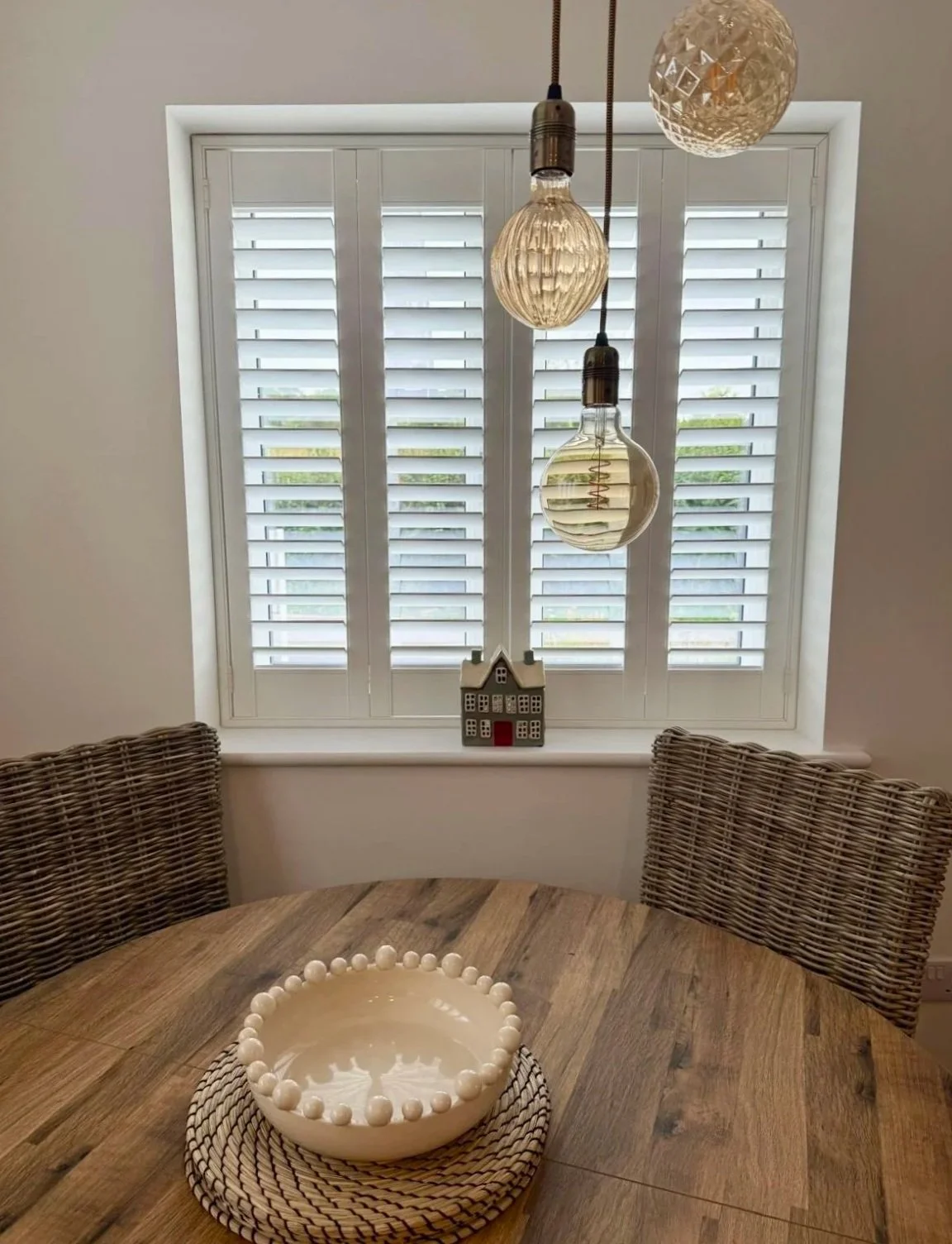 Dining room with wooden table, wicker chairs, a window with white shutters, hanging light bulbs, a decorative house on the windowsill, and a ceramic bowl with a beaded border in the center of the table.