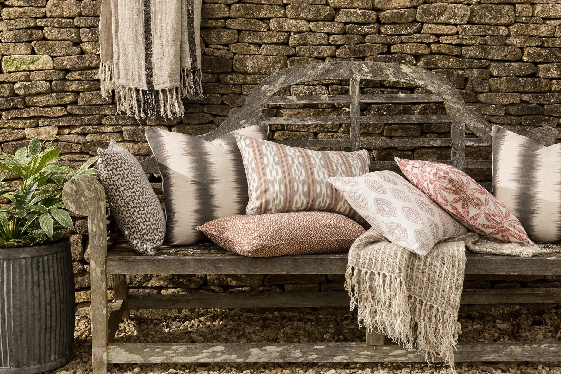 A rustic wooden bench decorated with multiple patterned cushions and a cozy blanket, set against a stone wall with a hanging textile and a potted plant nearby.