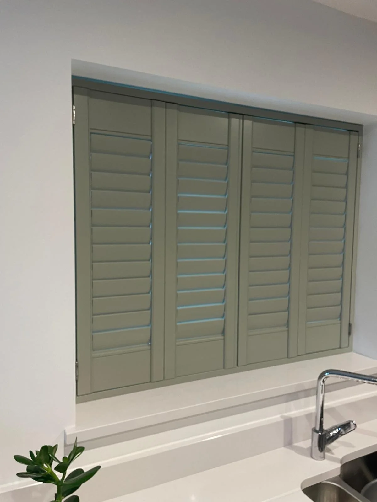 A view of closed gray window shutters on a kitchen wall, with a white countertop and a silver faucet in the foreground, and a small green plant on the left side.