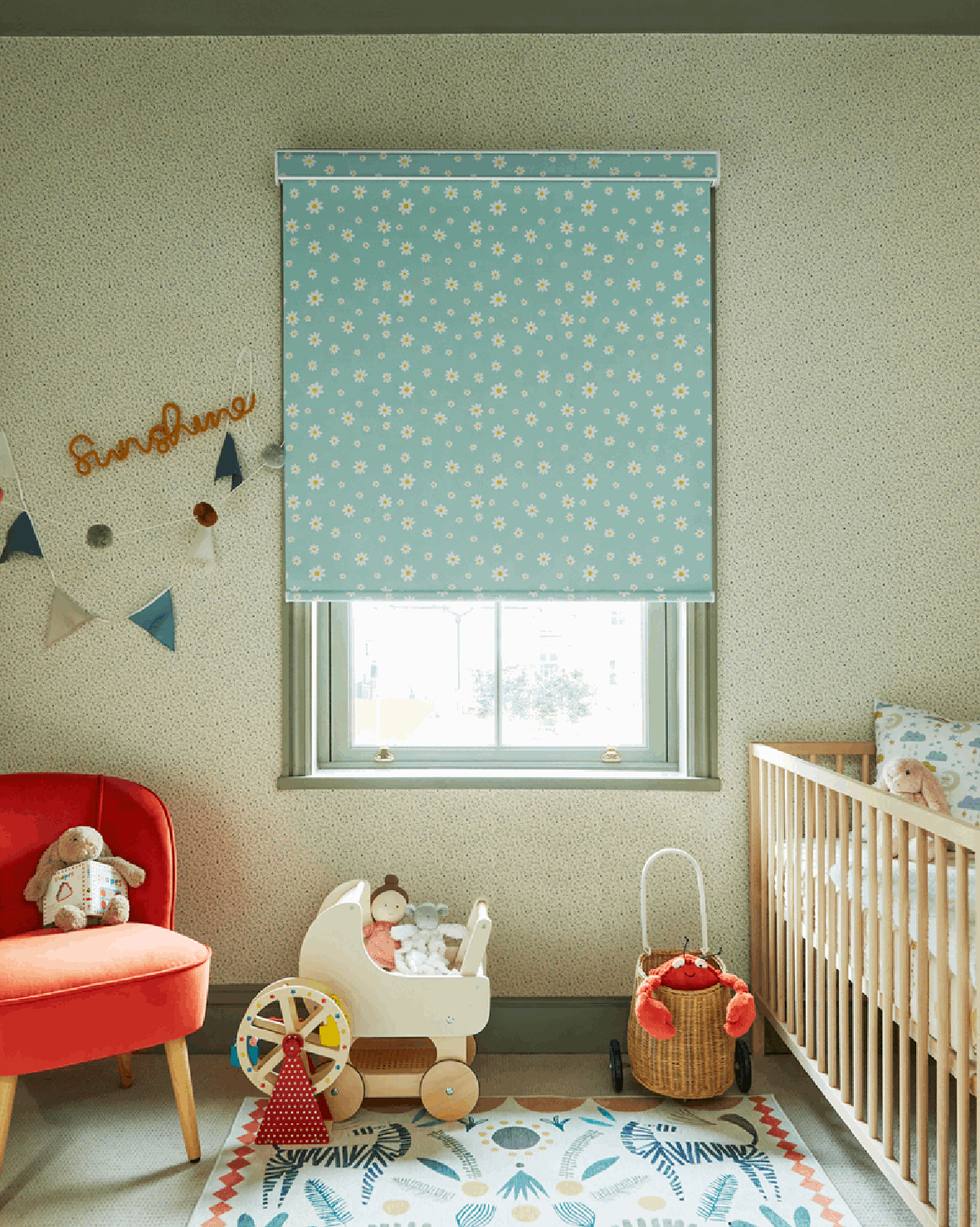 A cozy baby nursery with a window covered by a blue flower-patterned roller blind, a wooden crib with plush toys, a red armchair with stuffed animals, decorative wall bunting, and a colorful rug with botanical prints.