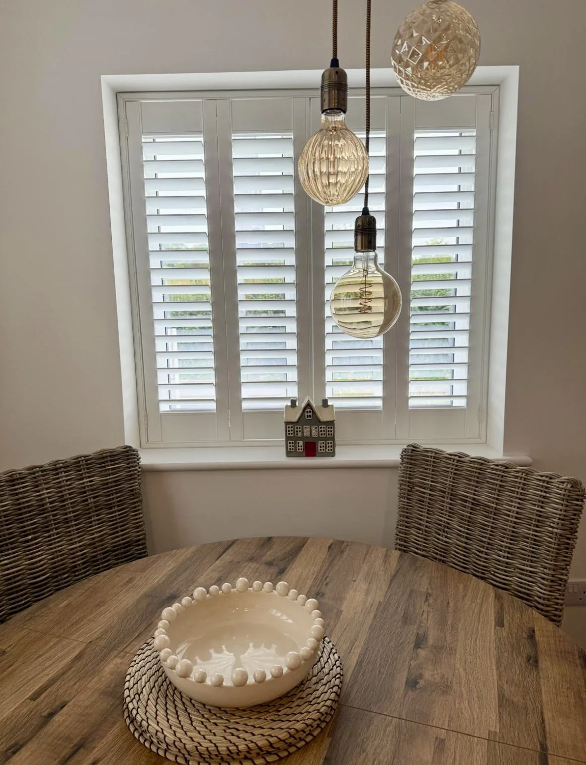 A round wooden dining table with a white ceramic bowl decorated with white balls, situated in front of a window with white shutters. Hanging pendant lights are above the table, and there is a small house-shaped decoration on the window sill.