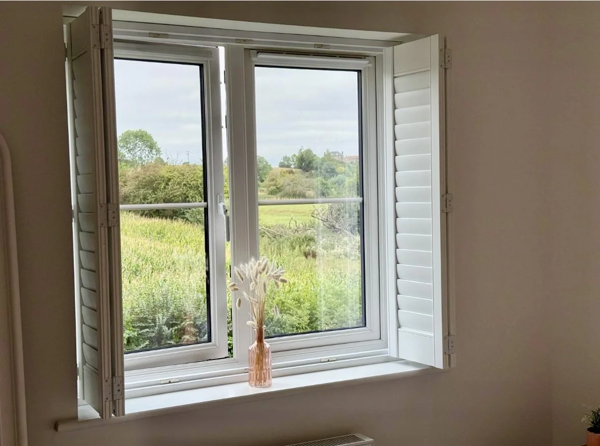 View through a window overlooking a green landscape with trees and a cloudy sky, with a pink vase and dried flowers on the windowsill.