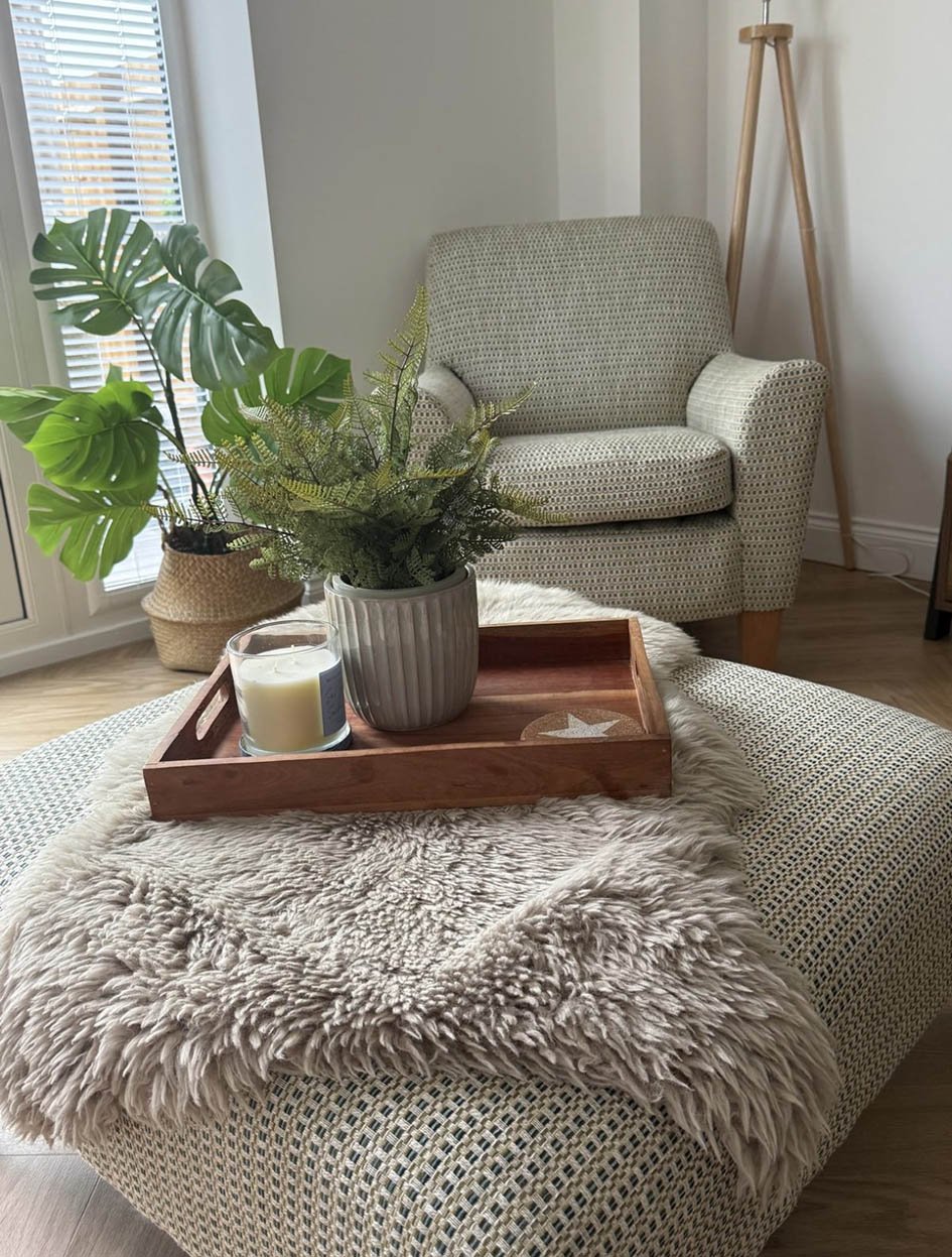 Living room with a patterned armchair, a large potted monstera plant, a wooden floor lamp, and a small table with a cozy faux fur blanket, a candle, and a plant in a ribbed pot on a wooden tray.