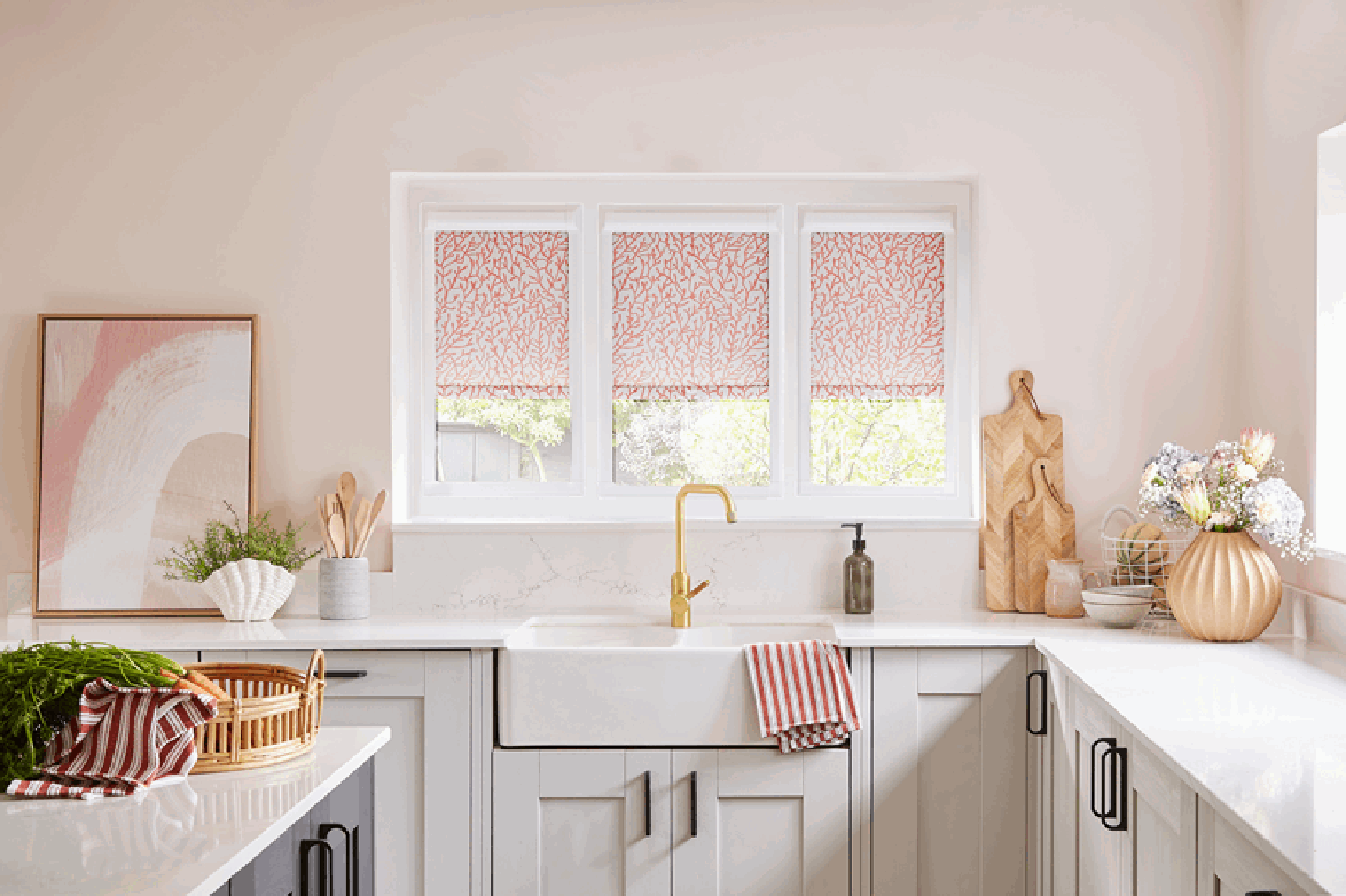 A bright, modern kitchen with white cabinets, a farmhouse sink with a gold faucet, and a window with pink patterned shades. Decor includes baking utensils, a large copper vase with flowers, and wooden cutting boards.