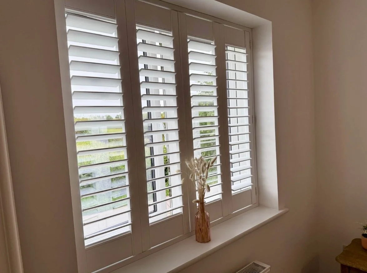 A window with white plantation shutters, decorated with a brown vase holding dried pampas grass on the windowsill.