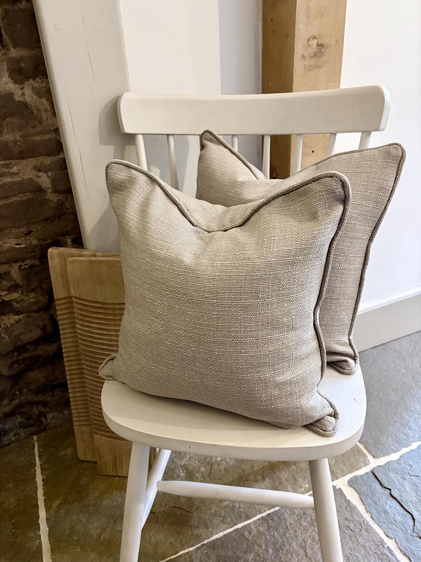 A white wooden chair with two beige textured pillows resting on it, positioned against a wall with a rustic brick accent.