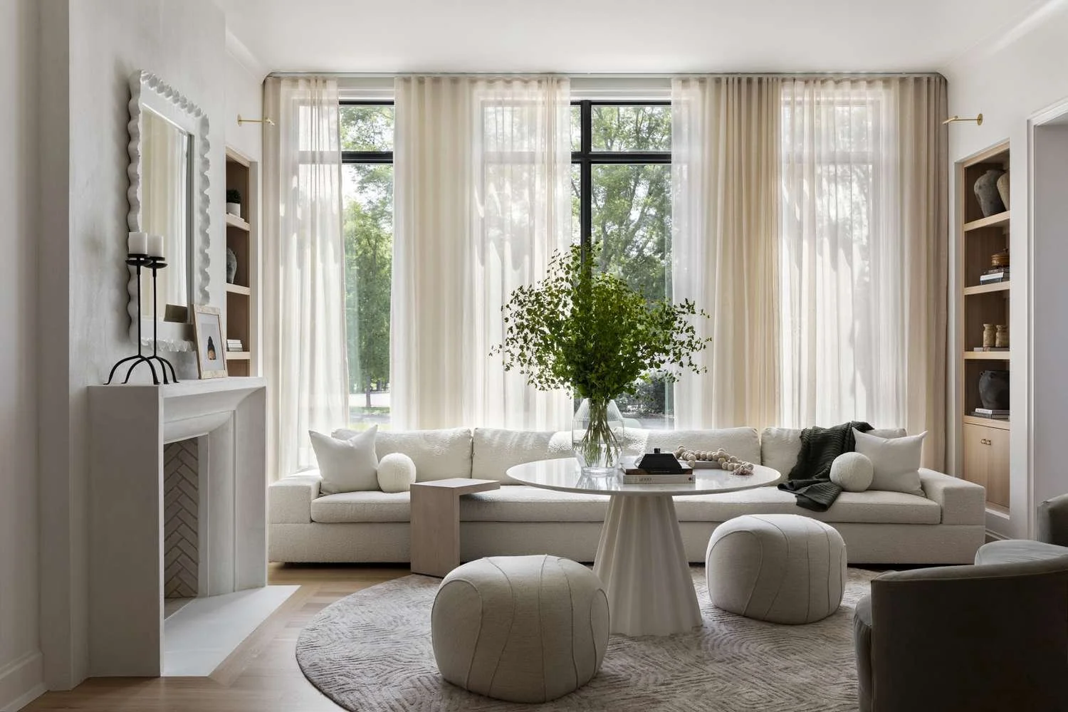 Bright living room with large windows, white sofa with pillows, a round coffee table, and decorative shelves with vases and art.