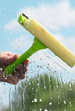 Person cleaning glass window with a green sponge and a yellow squeegee, water splashing.