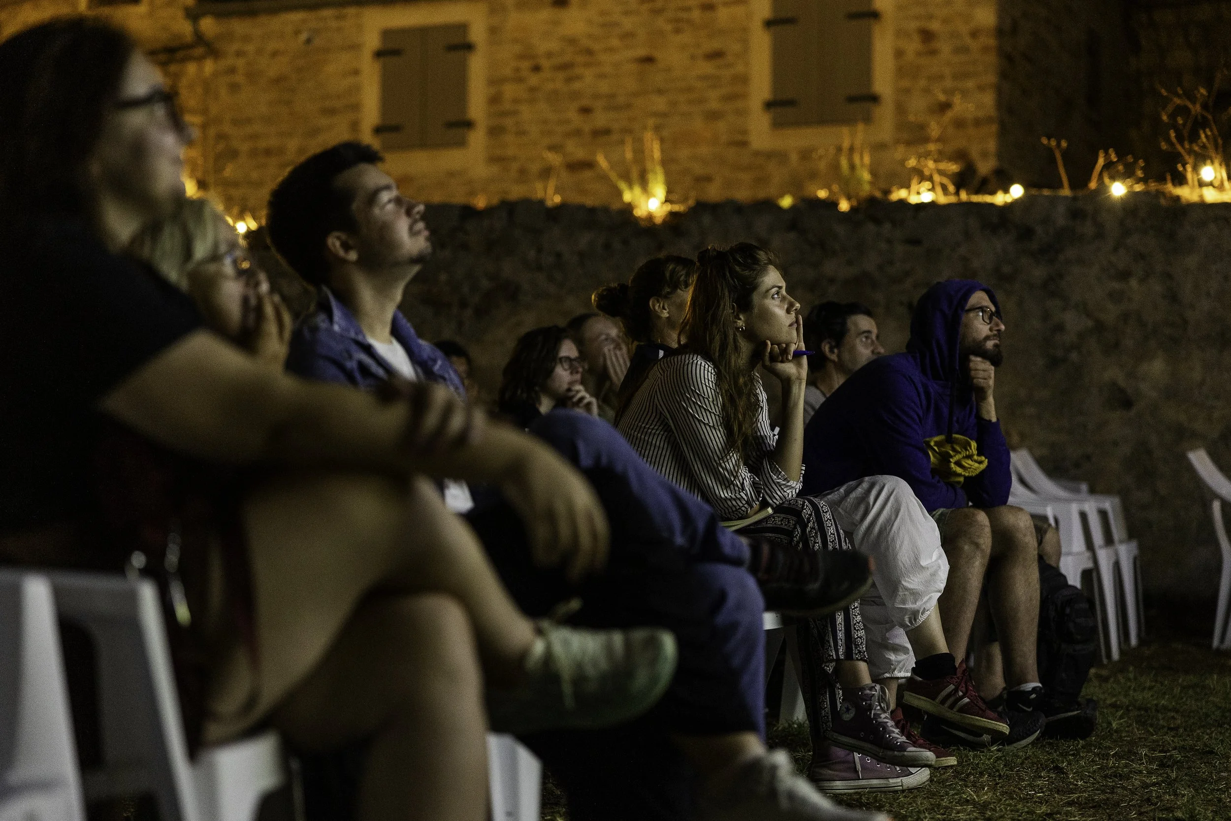A group of people sitting outdoors at night, watching a screen, with a brick building illuminated in the background.