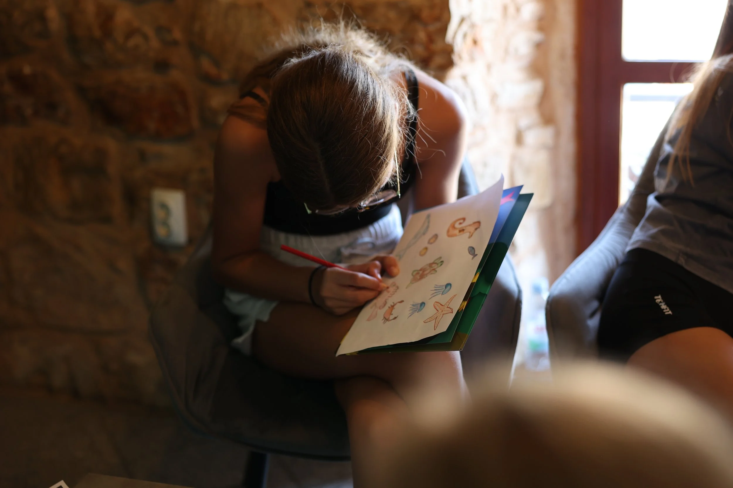 A person sitting on a chair, looking down and drawing or coloring pictures of sea creatures in a notebook. The person has long hair, is wearing a tank top, and is in a room with a brick wall and a window.