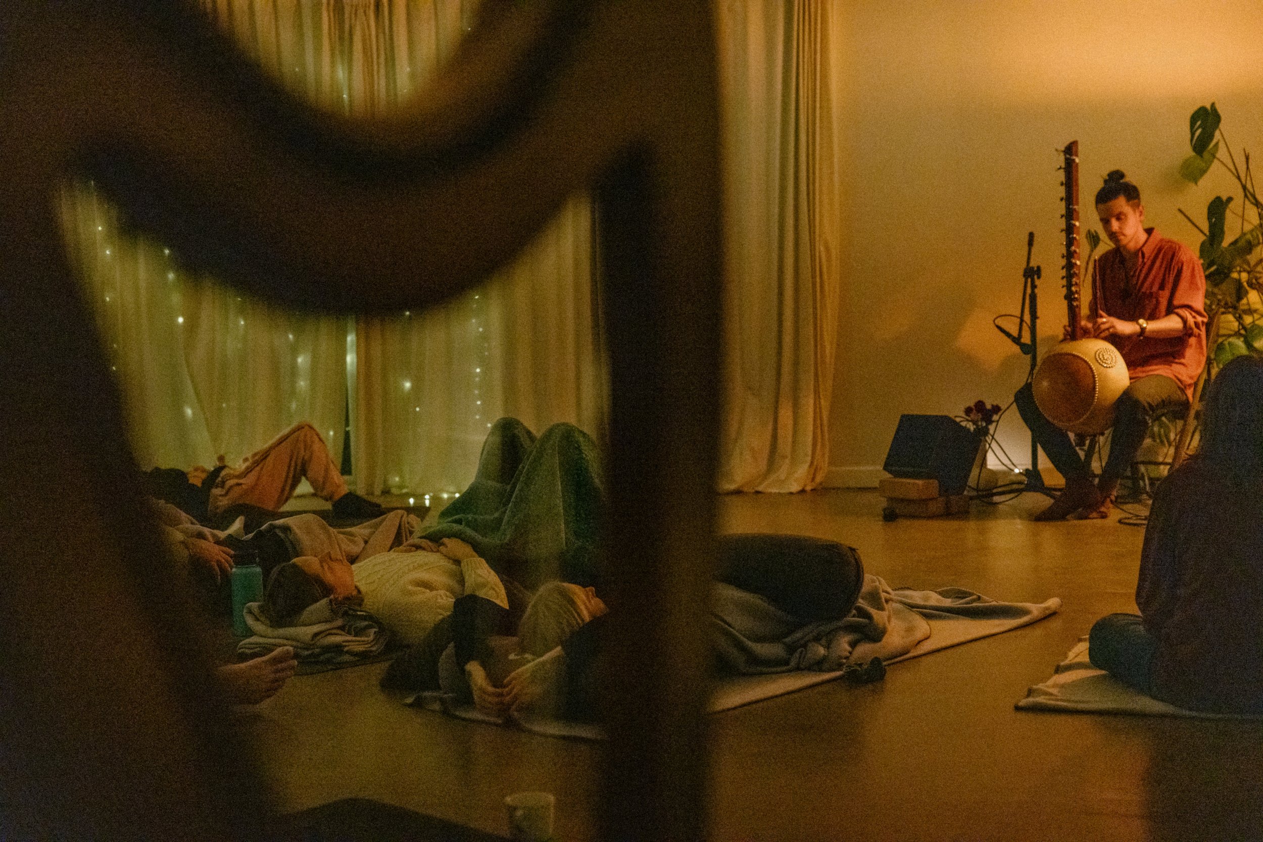 A musical performance in a dimly lit room with people lying on the floor, Josh Doughty sitting on a chair playing a the kora instrument, and a curtain with string lights in the background. at yoga sesstion