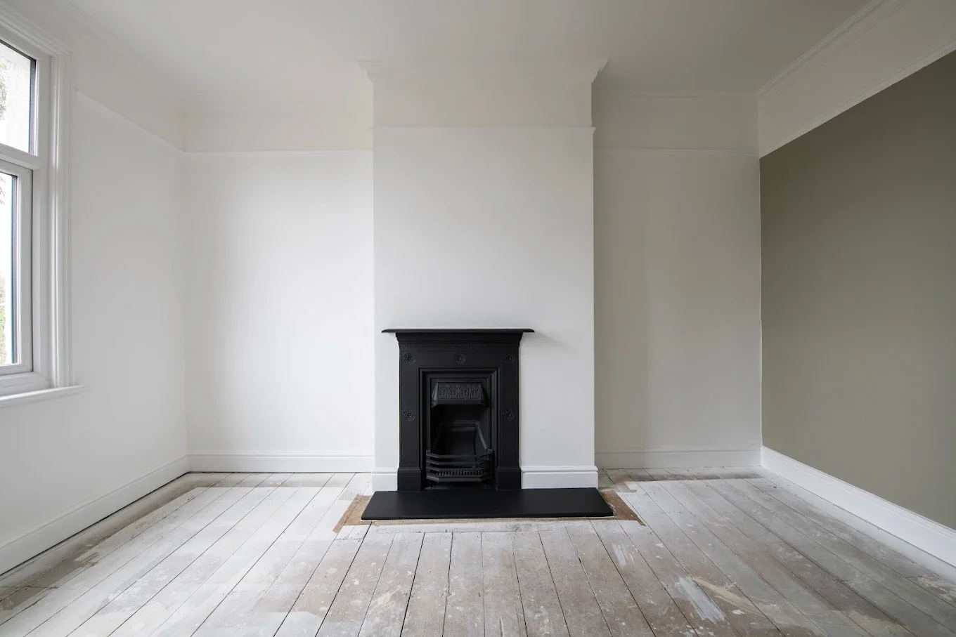 Empty room with white walls, a black fireplace in the center, a window on the left, and unfinished wooden floors.