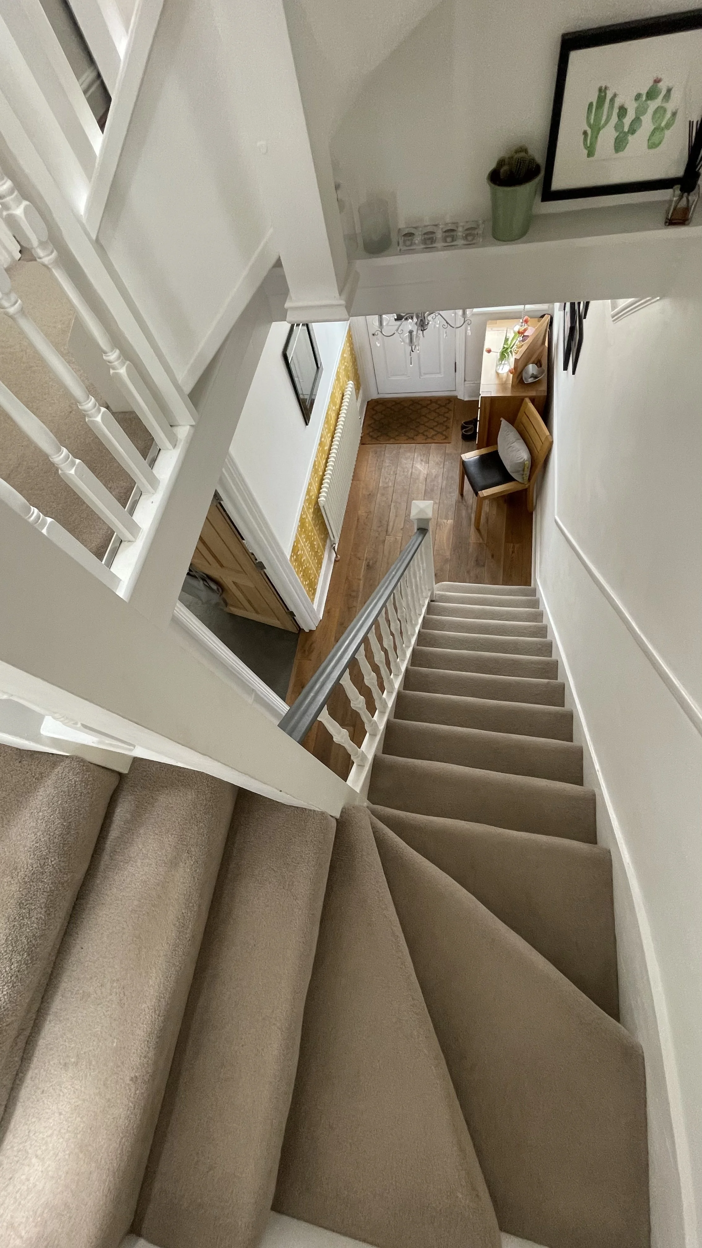 View looking down a staircase into an entryway with hardwood floors, a front door, a small table with a plant on it, a picture on the wall, and a chair with a pillow.
