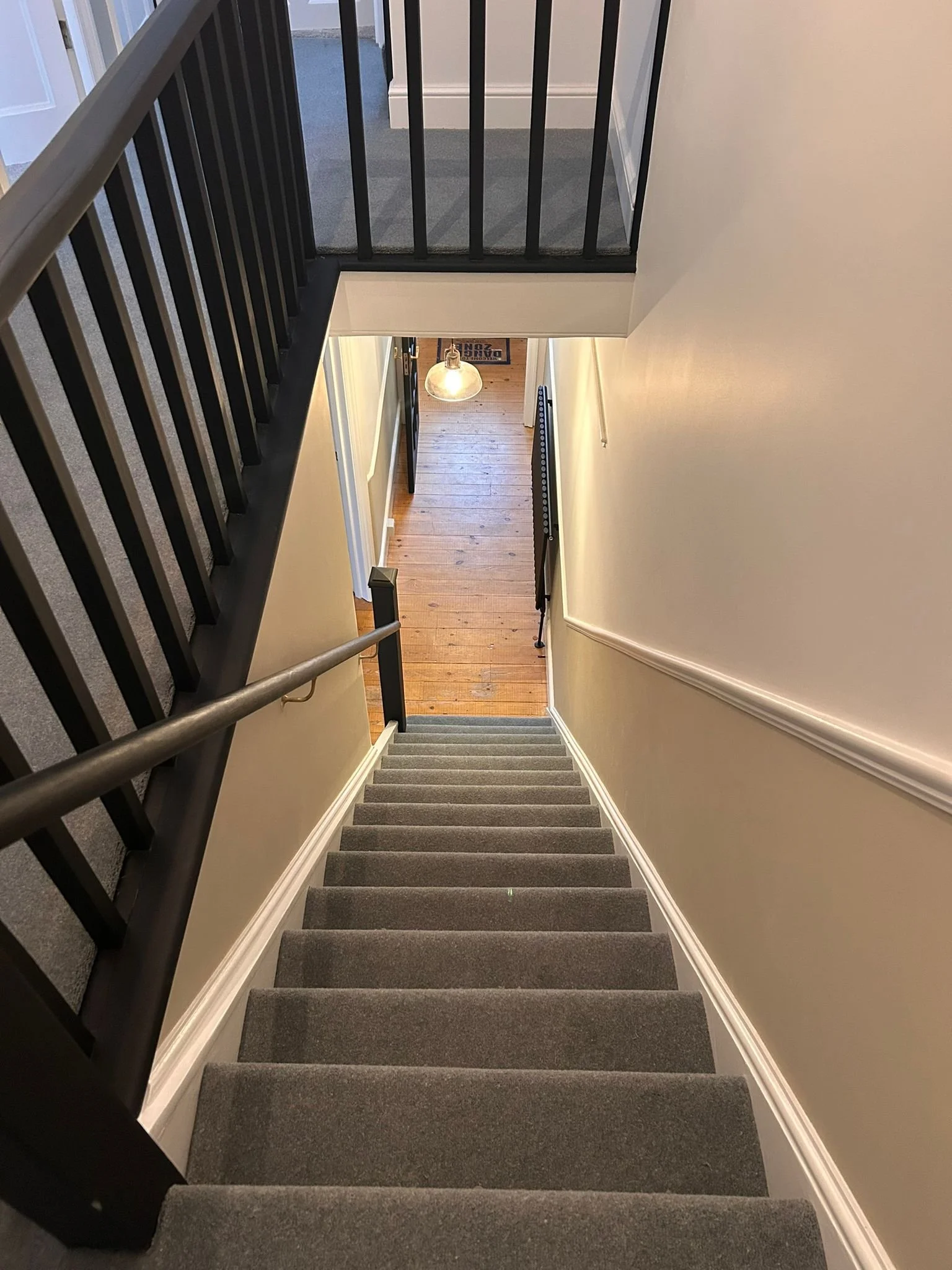 A view looking down a staircase with gray carpet, black railing on the left, white walls on the right, and a glimpse of a hallway with wooden floors and a hanging light fixture.