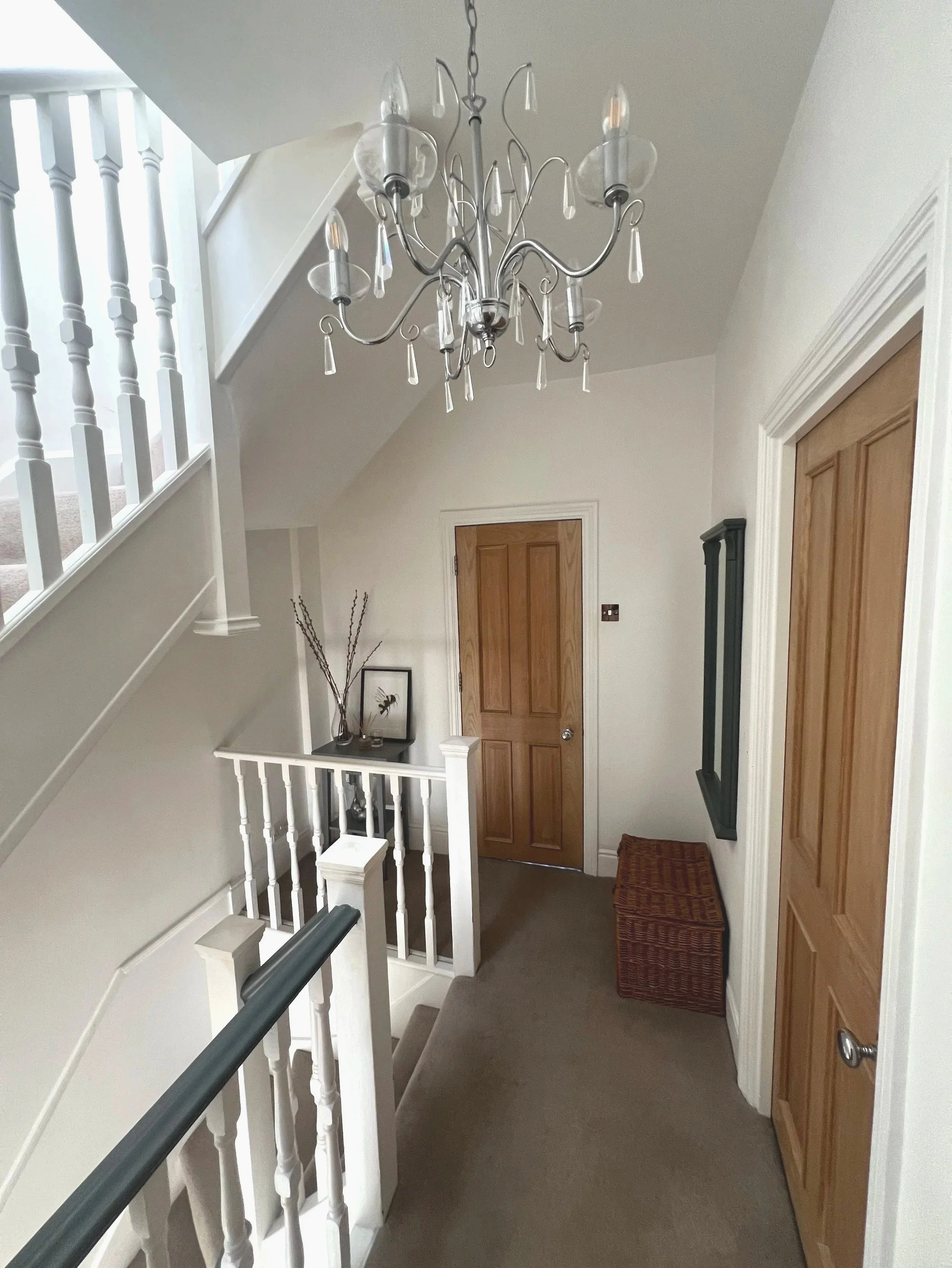 Interior view of a house hallway with a staircase, a wooden door, a chandelier overhead, and decorative items on a small table.