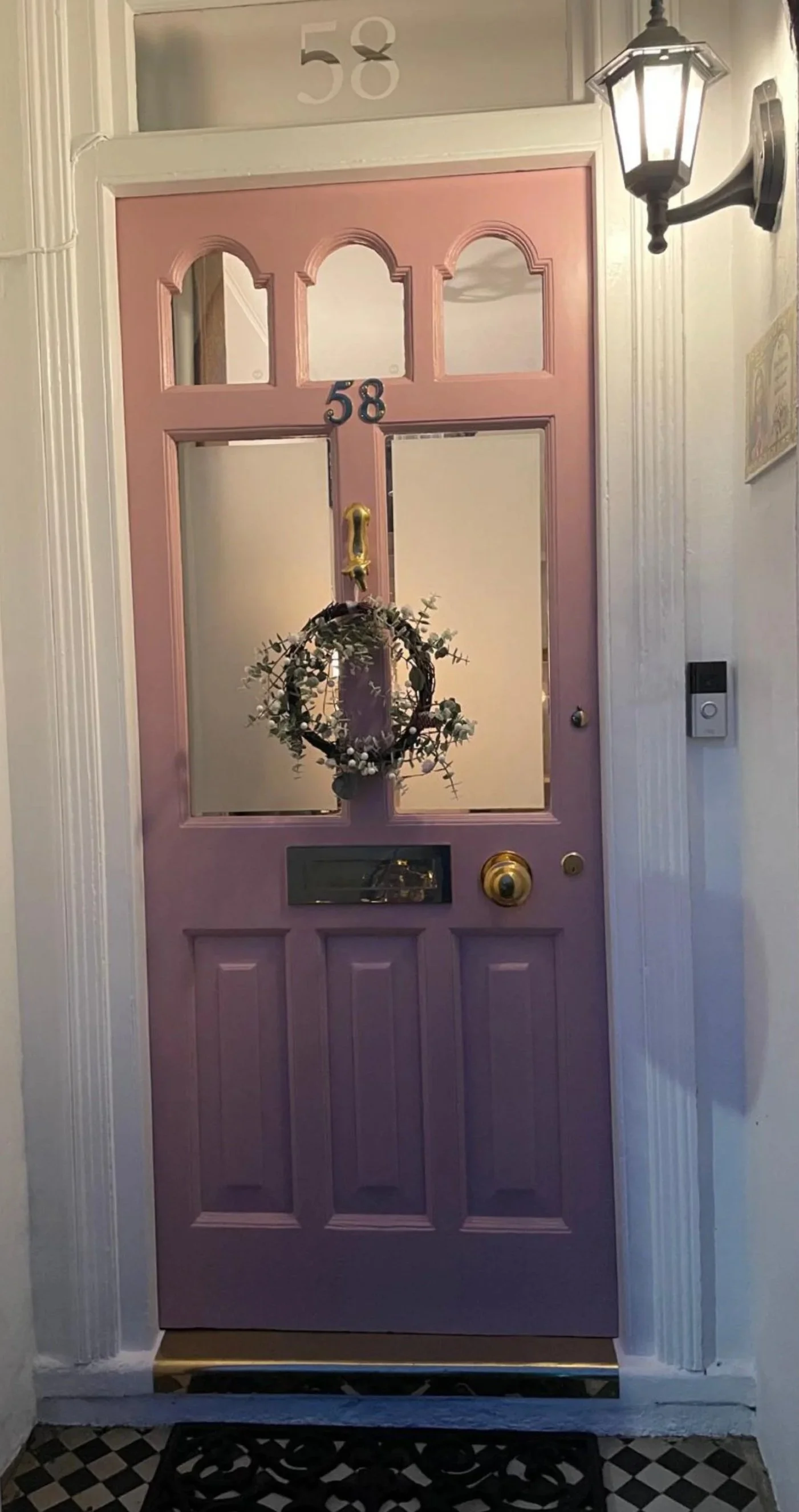 A pink front door with four small windows at the top, a decorative wreath in the center with white accents, and a gold door handle and mail slot. The house number '58' is displayed above the door and on the window above. There is a black lantern-style light fixture on the wall to the right of the door and a doorbell with a camera on the wall beside it. The floor has a black and white checkered pattern.