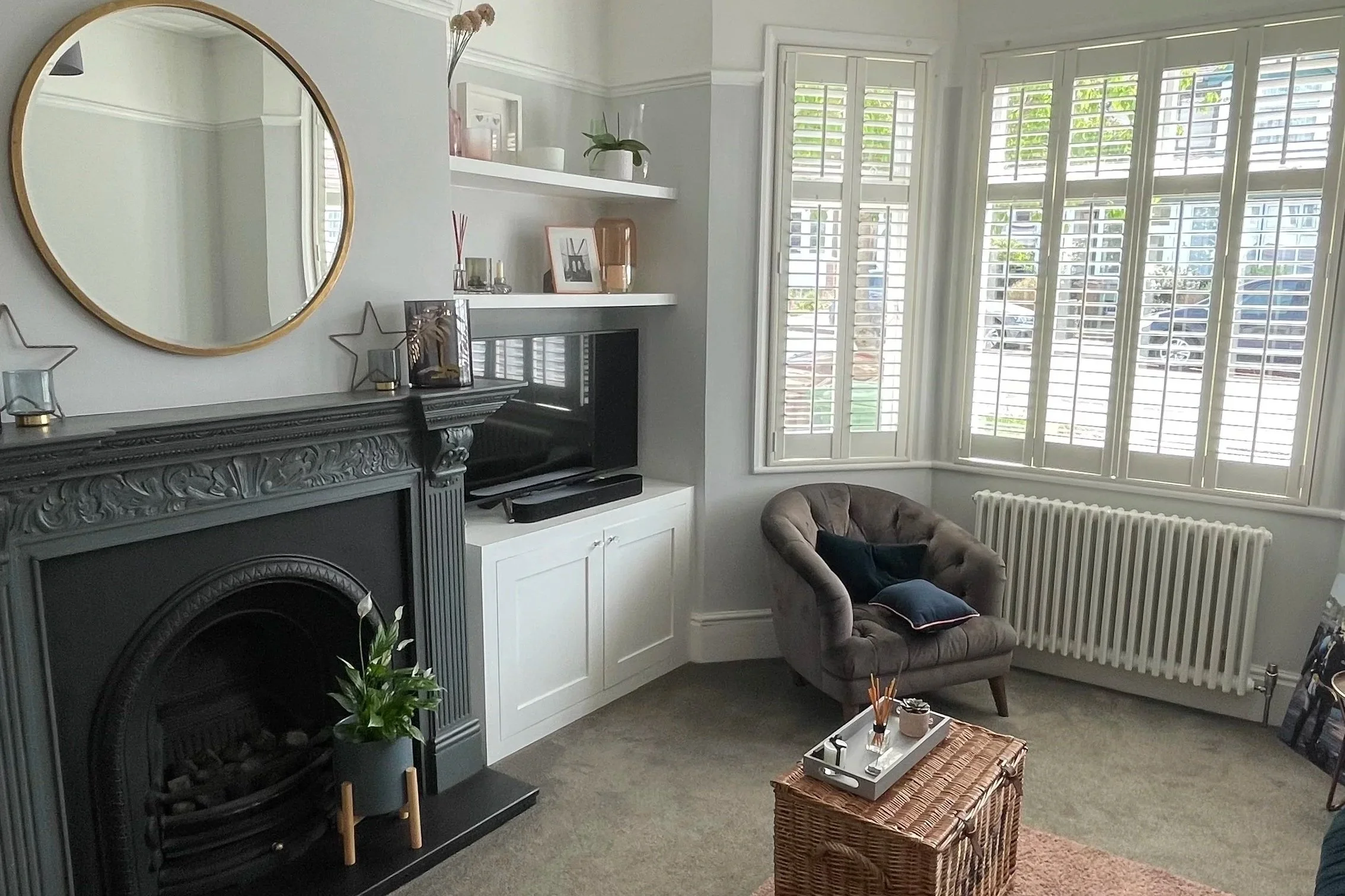 Living room with a black ornate fireplace, a large round mirror above it, a potted plant on the hearth, a flat-screen TV on a white cabinet, a gray tufted armchair with black and pink pillows, a wicker coffee table with a tray, tall window shutters allowing natural light, white shelves with decorative items, and a radiator below the window.