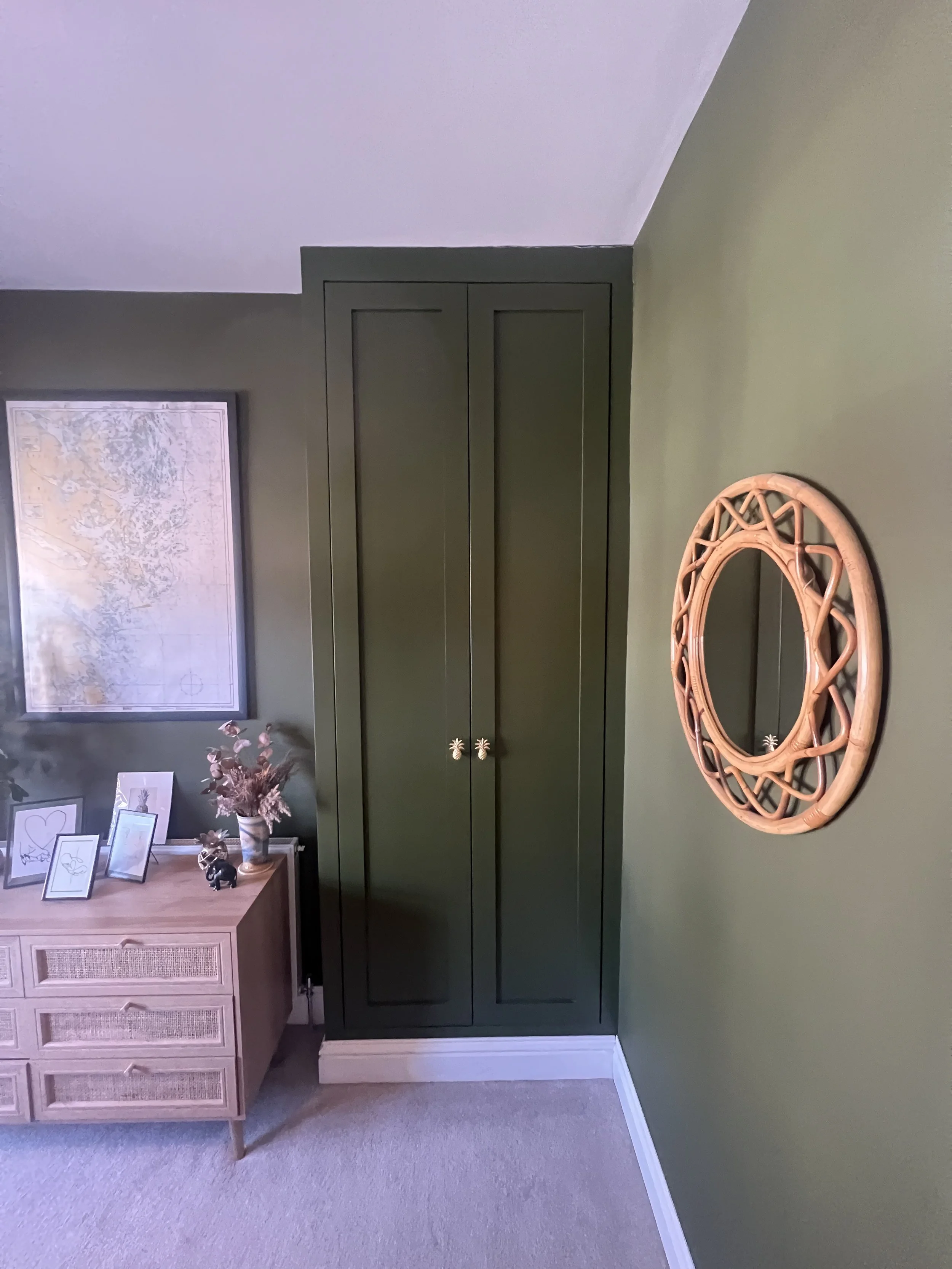 A green wardrobe with double doors, a wooden mirror frame on the wall, and a wooden dresser with framed photos and decor, in a room with green walls and beige carpet.