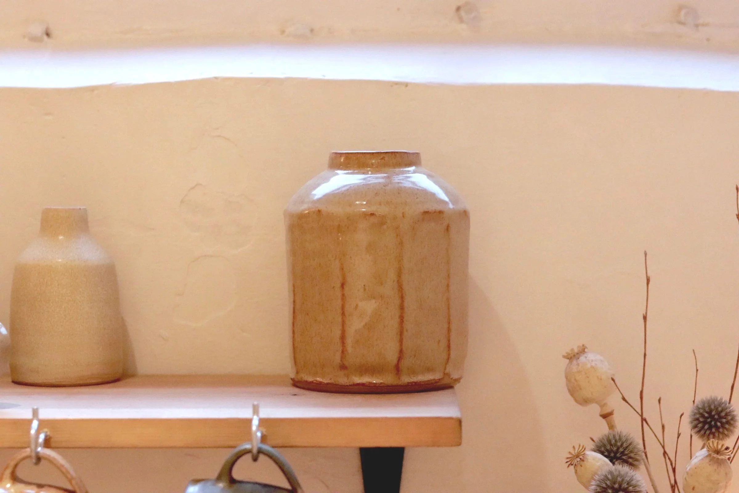Ceramic vases on a wooden shelf against a yellow wall, with dried flowers nearby.